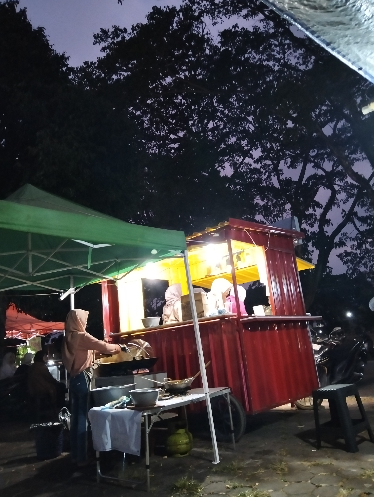 The image depicts a food stall operating at dusk in Magelang, Indonesia. A person, wearing a light brown hijab and a light brown hoodie, is actively engaged in cooking at a wok station located within the brightly lit, red food stall. In front of the stall, a foldable table is set up with various cooking utensils, including bowls and a gas cylinder. Another person, partially visible and wearing a white hijab, is also present at the stall. The stall is partially covered by a green canopy, and a smaller red canopy is visible in the background. The scene is illuminated by artificial lights from the stall, contrasting with the darkening twilight sky and the silhouettes of large trees overhead. The ground appears to be paved with interlocking stones and is wet in places. The overall atmosphere suggests a street food vendor at work in an outdoor urban setting.