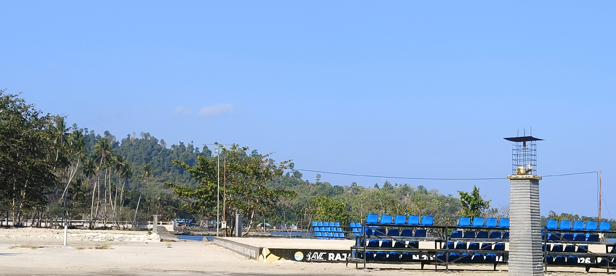 The image depicts a coastal scene in Waisai, Indonesia. In the foreground, a light-colored, sandy area leads to a concrete structure with a sign that reads "SAVE RAJA". To the right of this structure are rows of blue stadium-style seats, some of which are empty. Behind the seating area, a tall, grey, pillar-like structure stands, topped with a metallic cage-like enclosure containing some objects. A clear blue sky dominates the upper portion of the image. In the middle ground, palm trees and other lush green vegetation line the coast, with a body of water visible beyond them. Further in the background, a densely forested hillside rises. The overall impression is a scenic, potentially recreational area on a clear, sunny day. There are no people visible in the image.