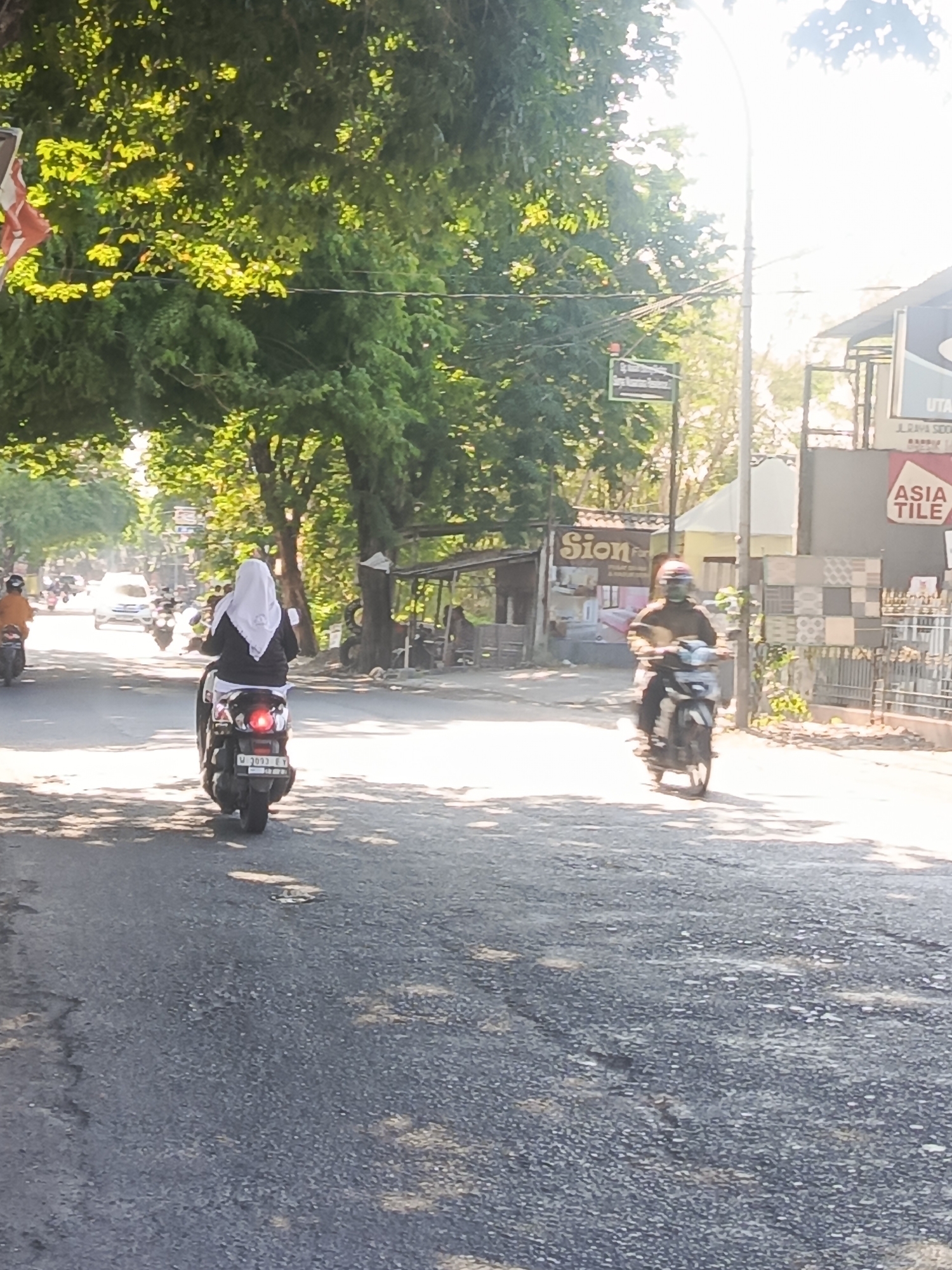 A daytime scene depicts a street in Sidowungu, Indonesia, with two individuals riding motorcycles. The foreground shows a gray asphalt road with patches of sunlight and shadows cast by overhead trees. On the left, a motorcycle is moving away from the camera, ridden by a person wearing a white hijab and a dark jacket. The motorcycle has a license plate that reads "W 3033 EV". Further down the road, another motorcycle is approaching, with a rider wearing a helmet and a dark top. In the background, there are buildings, including one with a sign for "Sion" and another with "ASIA TILE" branding. Lush green trees line the sides of the street. The lighting suggests it is a sunny day, possibly late morning or early afternoon.