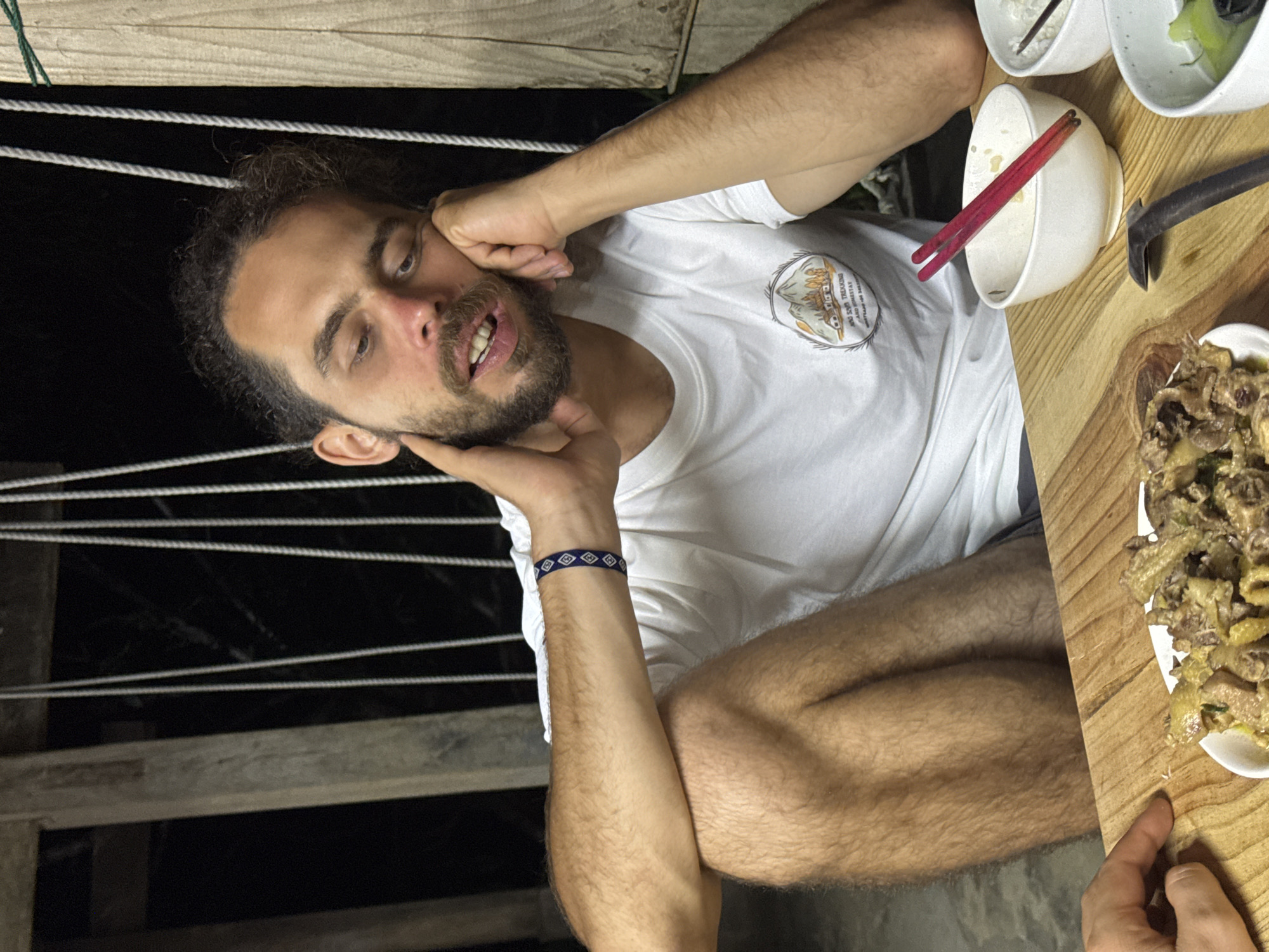 A man with a beard and a white t-shirt is sitting at a wooden table with food and bowls. The table is outside, near a wooden railing with ropes. The location is Pờ Ly Ngài Commune, Vietnam. The man has one hand on his face and is looking towards the camera. A plate of food, bowls, and chopsticks are on the table.