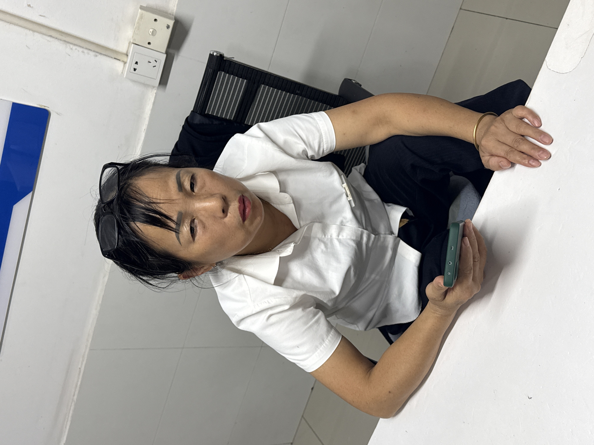 A woman is seated at a white table, leaning forward and looking at the camera. She is wearing a white button-down shirt, black pants, and sunglasses perched on top of her head. A gold bangle is visible on her left wrist. Her right hand is holding a green smartphone. The background shows a white tiled wall with electrical outlets. Behind her, a black chair with a mesh back is visible, and to her left, a portion of a blue and white sign can be seen. The setting appears to be indoors, possibly an office or waiting area, given the presence of the outlets and office chair. The lighting suggests it is daytime. The woman's expression is neutral.