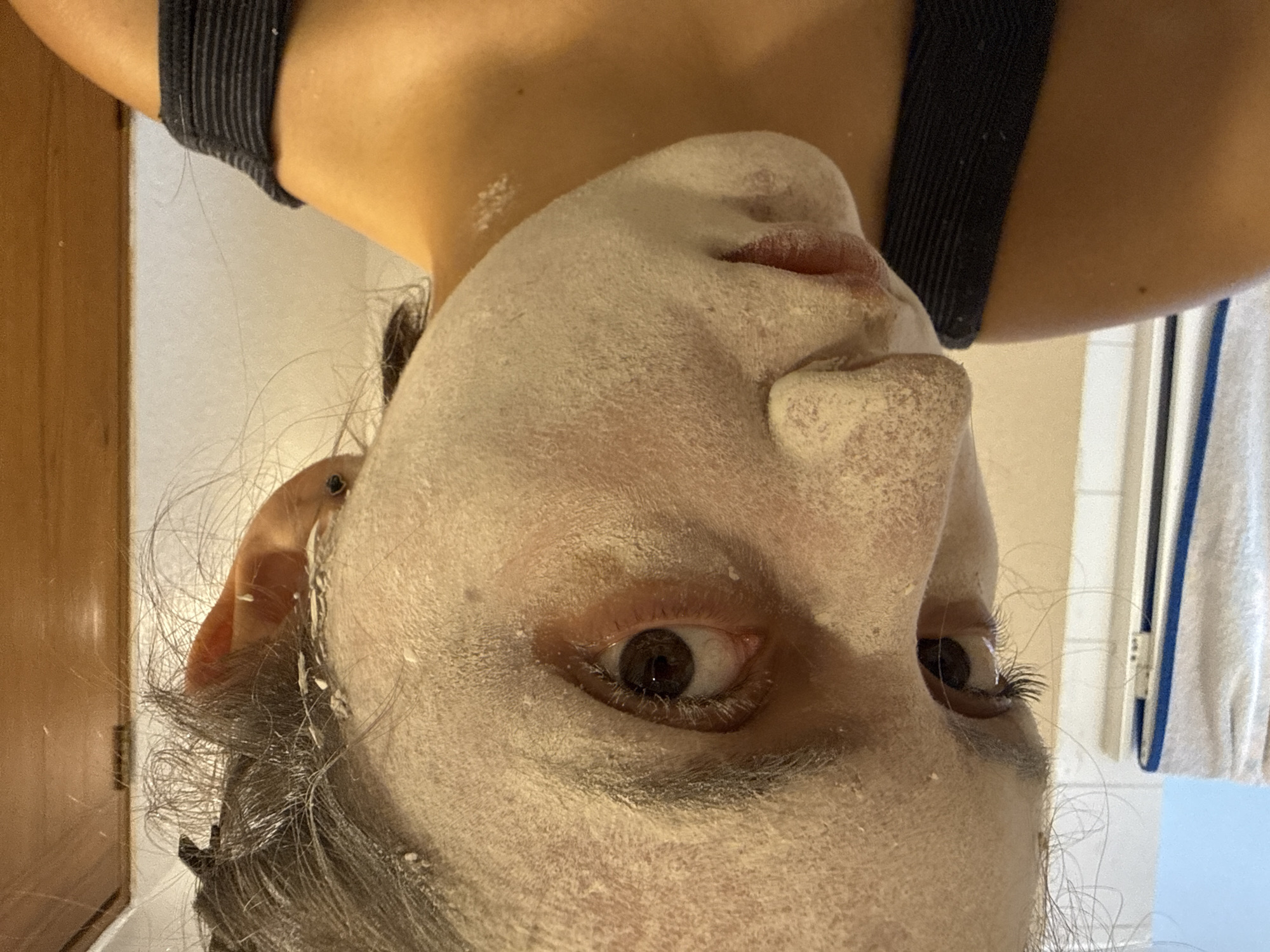 A person with a white facial mask is facing the camera in an upside-down orientation. They have brown eyes and graying hair. A black strap is visible on their shoulder. The background includes a wooden door and a towel rack with a towel. The location cannot be determined to be Mexico City, Mexico from the image.