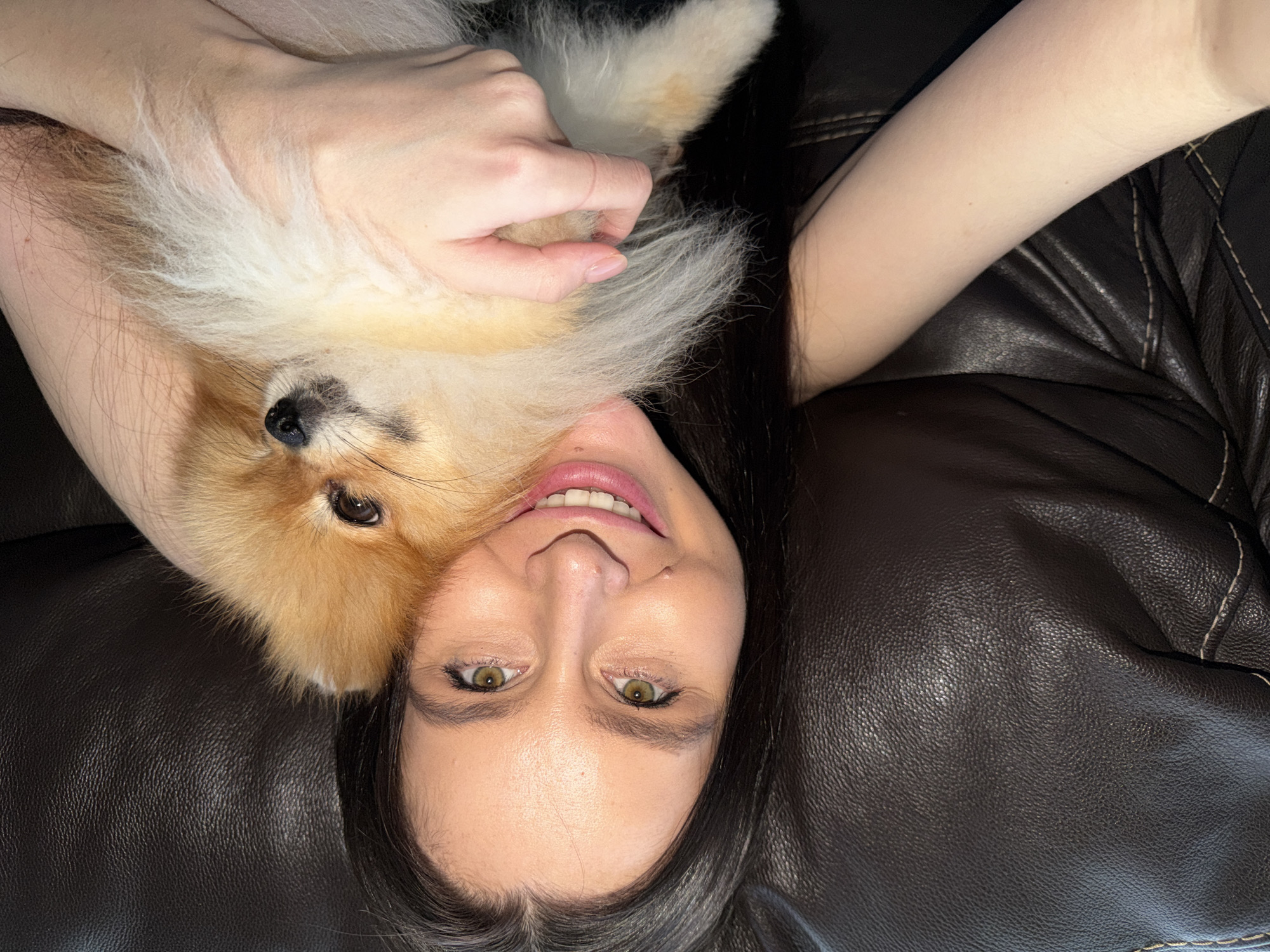 An overhead shot captures a heartwarming moment between a woman and her small, fluffy dog, lying together on a dark brown or black leather sofa. The woman, with long dark hair, fair complexion, and distinctive green eyes, is positioned at the bottom of the frame, looking upwards with a gentle, open-mouthed smile that reveals her teeth. Her expression radiates contentment and warmth.
Cuddled against her, likely on her chest or shoulder, is a small canine, possibly a Pomeranian, characterized by its soft, voluminous fur in shades of light brown, orange, and white. The dog's dark nose and eyes are visible, and it appears relaxed and comfortable. The woman's hand is gently placed on the dog's fur, emphasizing the affectionate bond between them.
The backdrop is a textured leather surface, indicating comfortable indoor furniture, possibly a sofa or armchair. The perspective is directly from above, providing a close-up and personal view of the interaction. The overall mood is one of peace, affection, and mutual companionship. No text is visible in the image. The scene is set in Khimki, Russia.