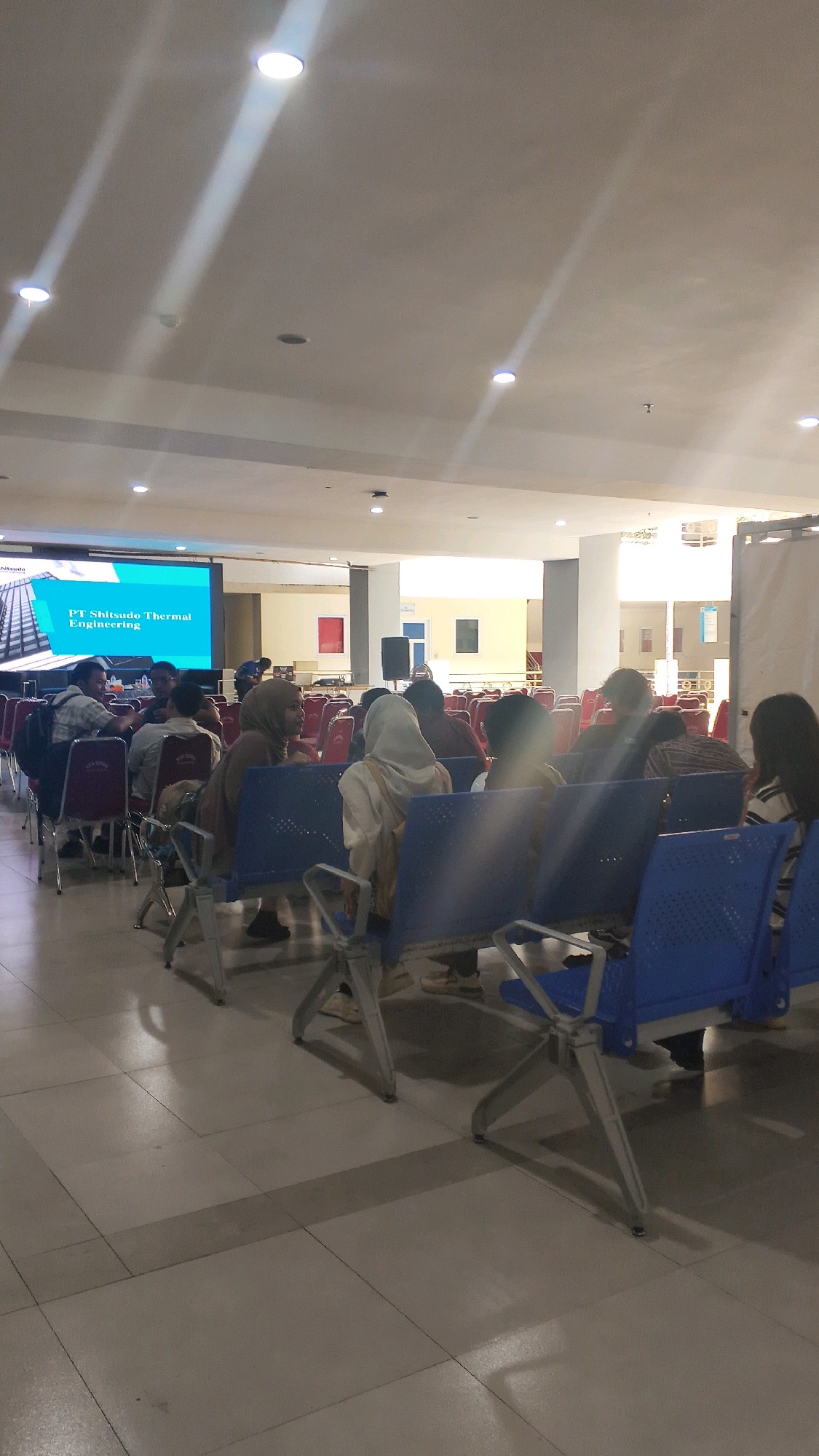 The image depicts an indoor scene, likely a conference room or waiting area, with several individuals seated on blue chairs. A large screen displays the text "PT Shitsudo Thermal Engineering". The room has a tiled floor and is illuminated by overhead lights. Some individuals are facing the screen, while others are engaged in conversation or looking away. The overall impression is of a formal gathering or presentation. The location is specified as Lohbener, Indonesia.