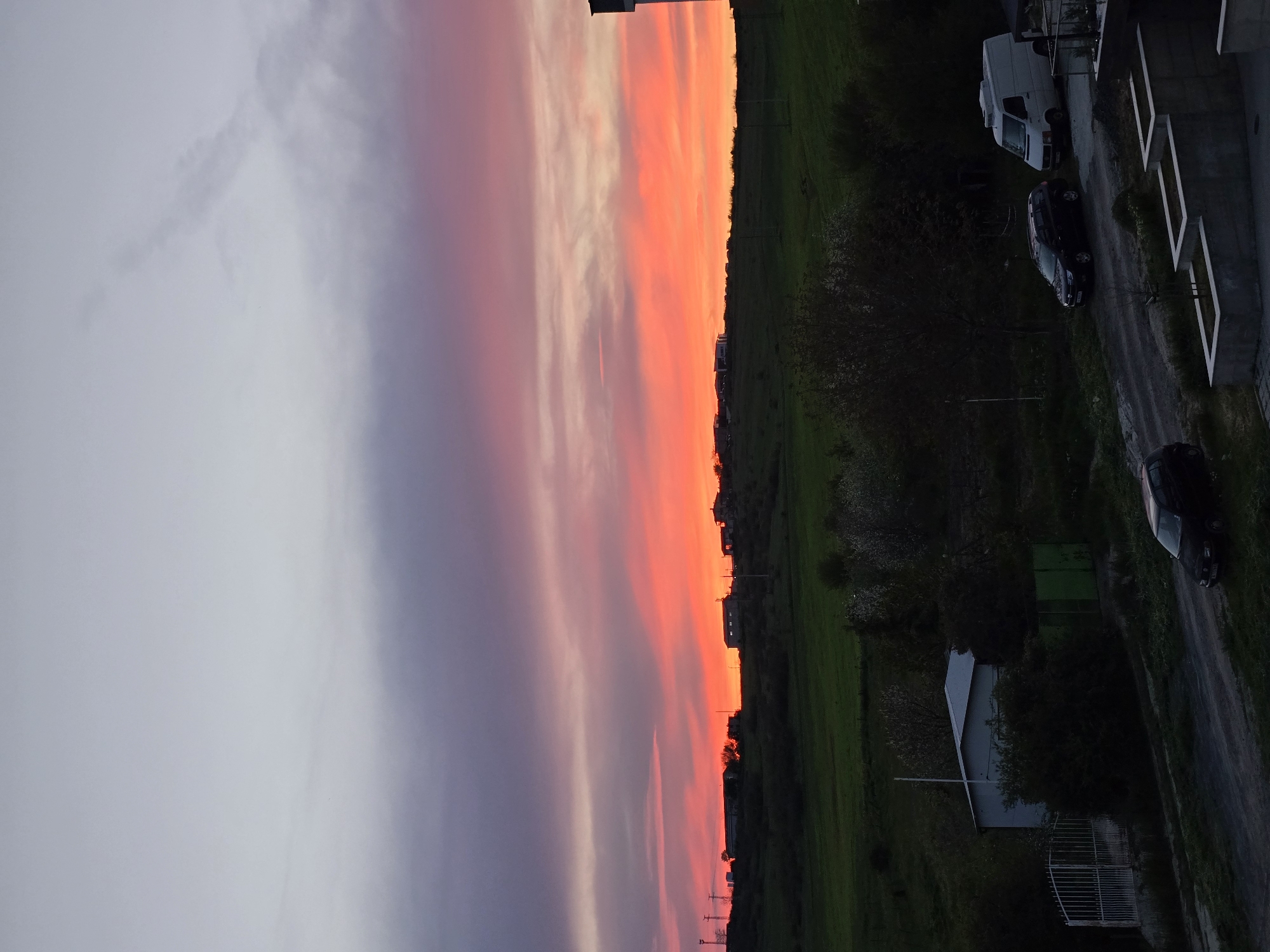 The image shows a landscape at dusk or dawn in Burgas, Bulgaria. The sky is a gradient of light gray to pink and orange, indicating either sunrise or sunset. Below the horizon, there is a dark green field bordering a street with parked vehicles: a white van, a dark-colored car, and another dark car. There's also a small white building with a fence. The overall impression is of a quiet residential area.