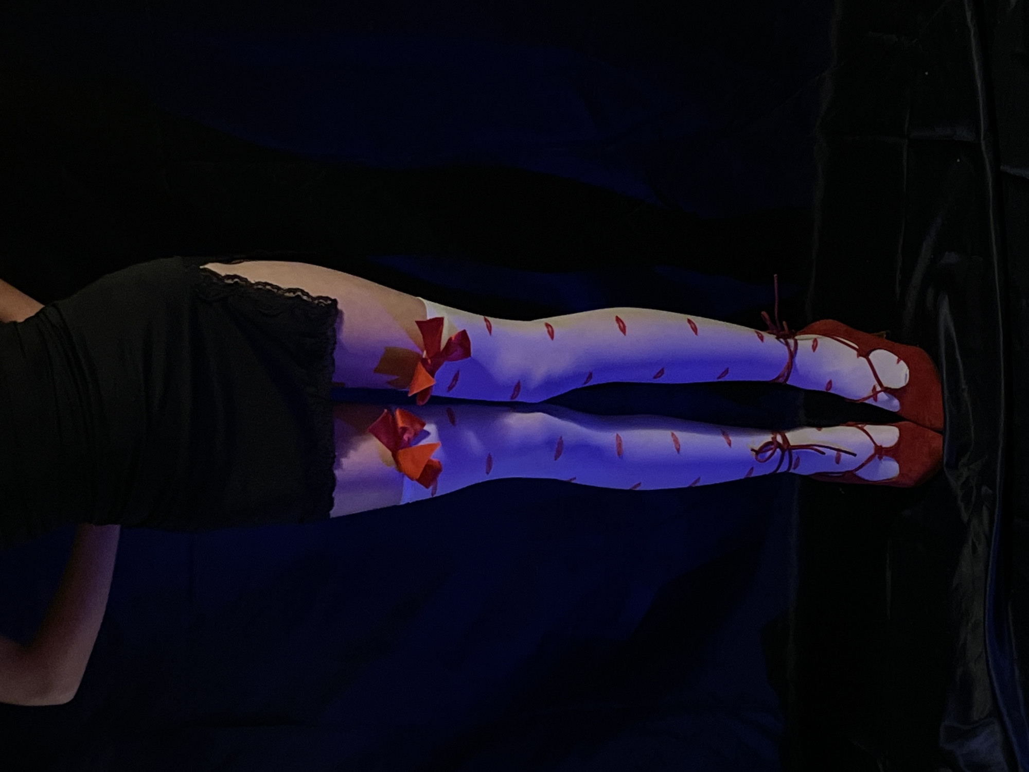 A person is lying down on a dark surface. They are wearing a black dress with lace trim, white stockings with red lip prints and red bows, and red shoes with laces. The lighting is dim, with a blueish hue. The background is a dark, possibly velvet, material. There is no indication of a specific location, including the city of Barcelona, Spain.