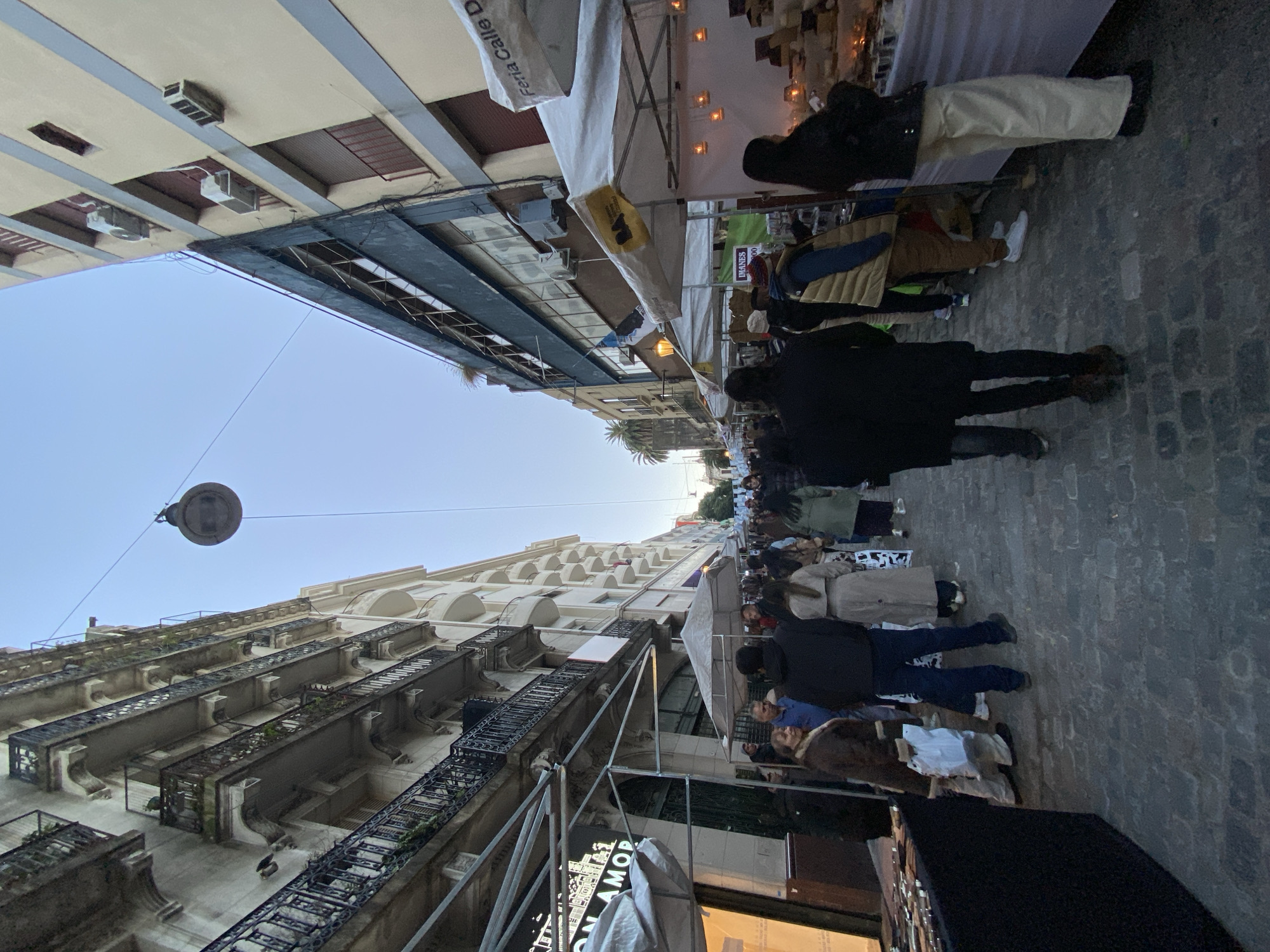 The image captures a street scene in Buenos Aires, Argentina. Cobblestone paving defines the pedestrian walkway, flanked by buildings with multiple floors and ornate iron balconies. Overhead, a street lamp is suspended by a cable against a clear sky. A bustling street market occupies the space, featuring vendor stalls covered with white canopies. Numerous people are present, milling about, presumably browsing the wares on display. The orientation of the image is tilted, providing an unconventional perspective on the urban setting.