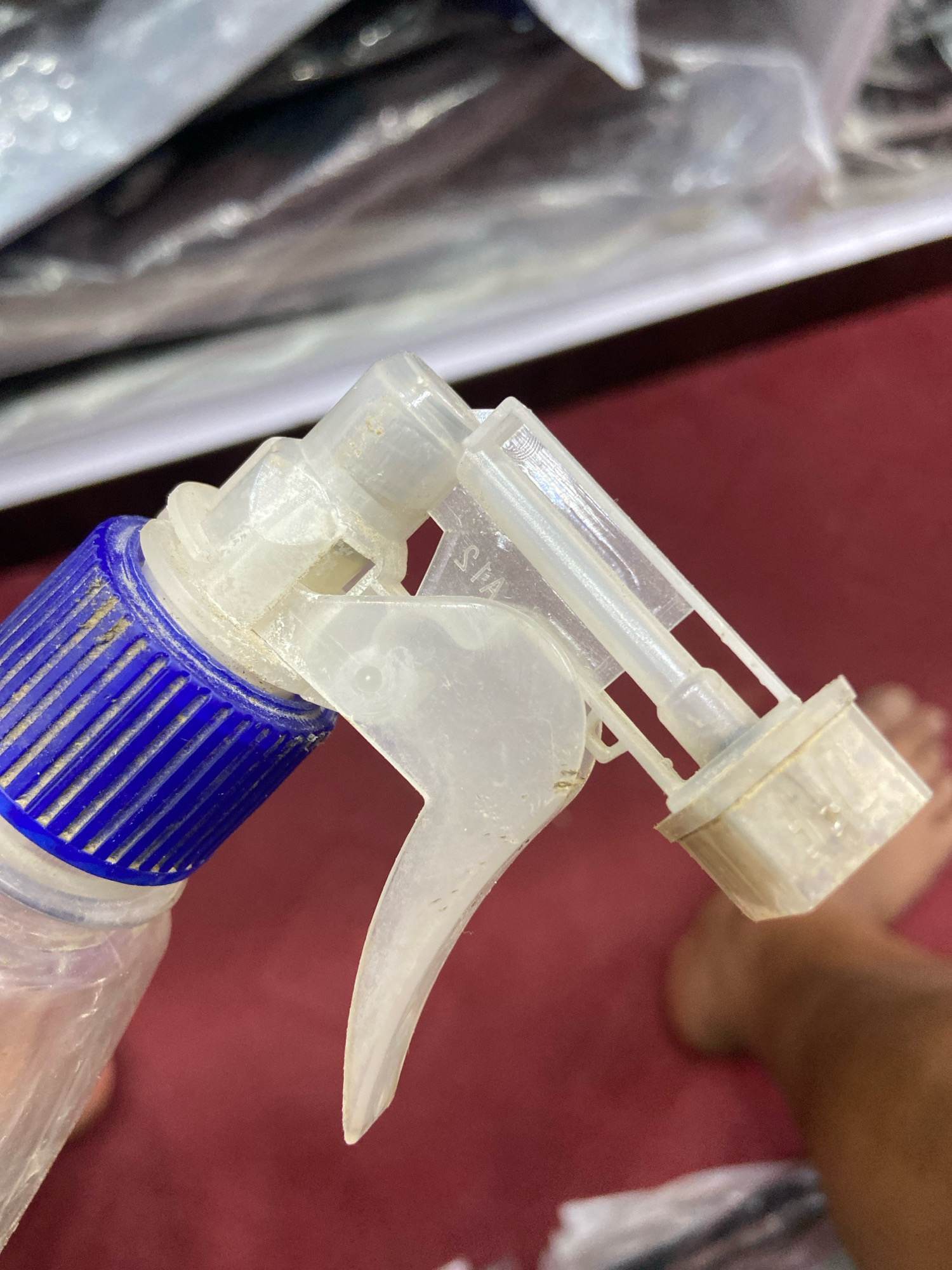 The image is a close-up of a used white plastic spray bottle nozzle with a blue ribbed cap. The nozzle is dusty, and some dried residue is visible around the trigger and the nozzle tip. The background is blurred, showing a reddish-brown surface and a person's bare foot with visible toes. The context of the image, specifying its location in Lahan, Nepal, is not visually discernible from the provided photograph itself.