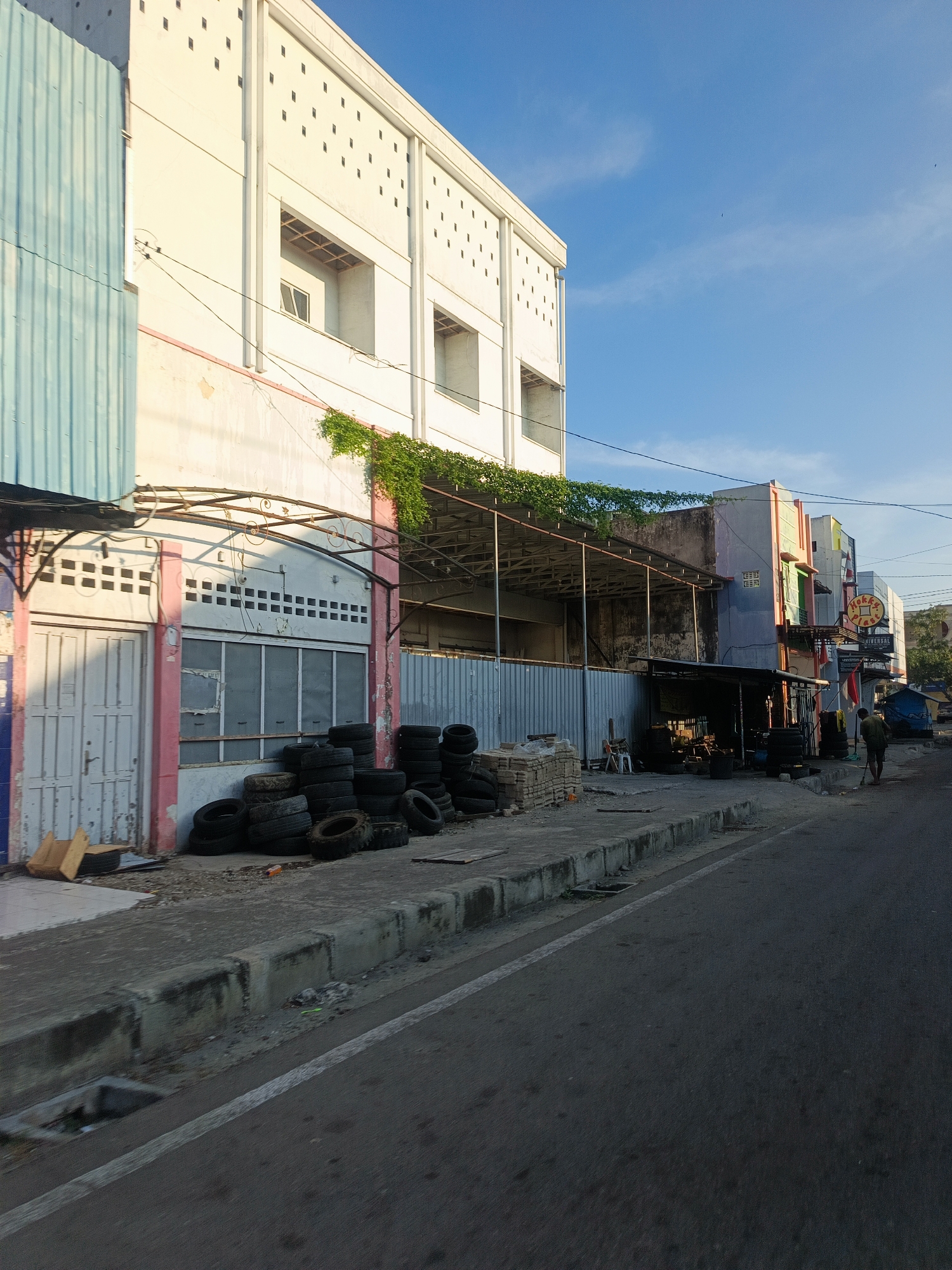 The image depicts a street scene in Gorontalo, Indonesia, likely a commercial area. The foreground shows a paved road with a white lane marker. Adjacent to the road is a concrete curb and sidewalk. Piled on the sidewalk are numerous used tires. Behind the tires, a building with a white facade and rows of small square openings is visible. To the left, a section of a building covered in corrugated metal, painted a muted blue, stands out. Further down the street, a series of other buildings line the road, some painted in various colors. A person is seen walking away from the camera on the sidewalk, carrying a broom. The sky is clear and blue. The scene appears to be captured during daylight, with soft lighting suggesting either early morning or late afternoon. The overall impression is one of a working-class or commercial district with a mix of old and newer structures.
