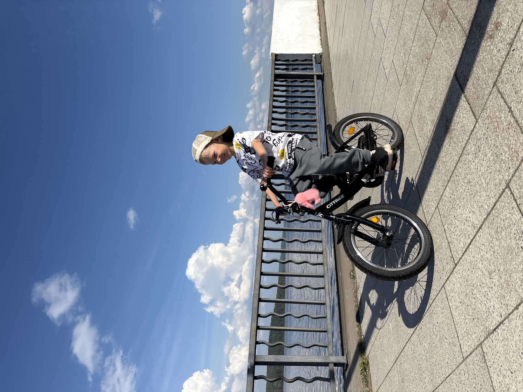 A young boy is riding a black bicycle on a paved walkway next to a body of water. The boy is wearing a beige baseball cap, a white t-shirt with black lettering, and grey pants. He is smiling and looking to his right. The bicycle has a pink fuzzy accessory attached to the handlebars. A metal railing separates the walkway from the water, and beyond the water is a tree-lined shore. The sky is blue with scattered white clouds. The scene appears to be in the city of Chaikovsky, Russia, based on the context provided.