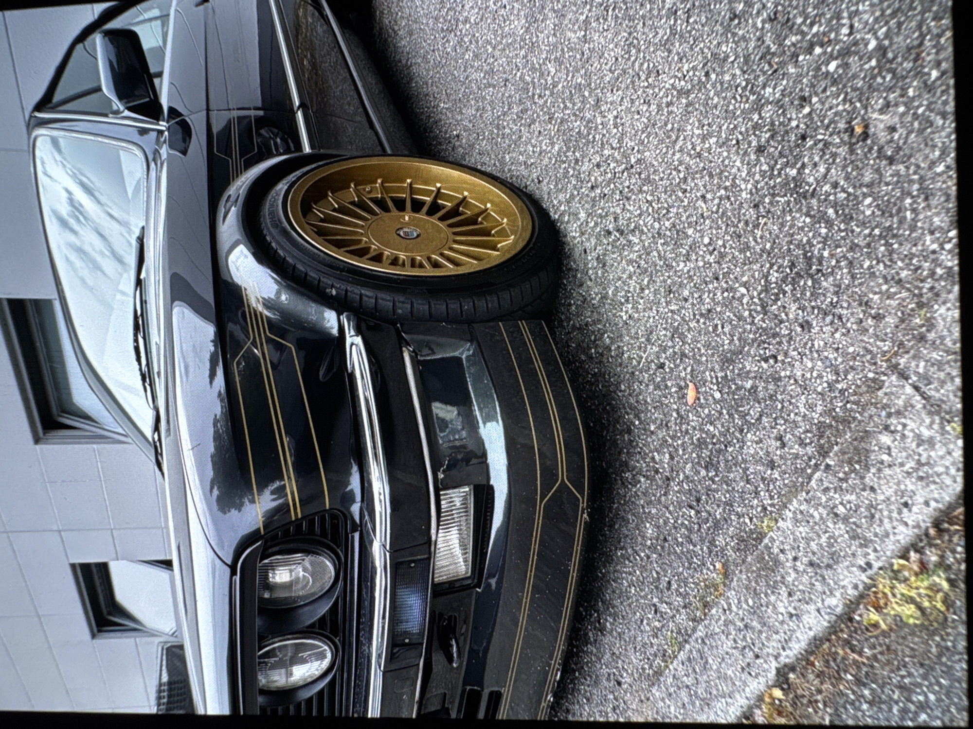 A detailed, low-angle, side-front perspective shows a classic black car with distinctive gold pinstriping parked outdoors. The car features prominent gold multi-spoke wheels fitted with black tires, on which the brand "HANKOOK" is visible. The front of the vehicle displays a dual round headlight design, a rectangular turn signal, and a black grille. Its glossy black paint reflects the surroundings, indicating a clean and well-maintained appearance.
The car is situated on an asphalt or tarmac surface, which extends across the ground and into the background on the right. To the left, a light-colored building with multiple rectangular windows is reflected in the car's windshield and side mirror. The scene suggests a daytime setting on a street or parking area in an urban or suburban environment. The car is stationary, and no people or specific activities are depicted. The image itself is oriented vertically, rotated 90 degrees clockwise from a typical horizontal view.