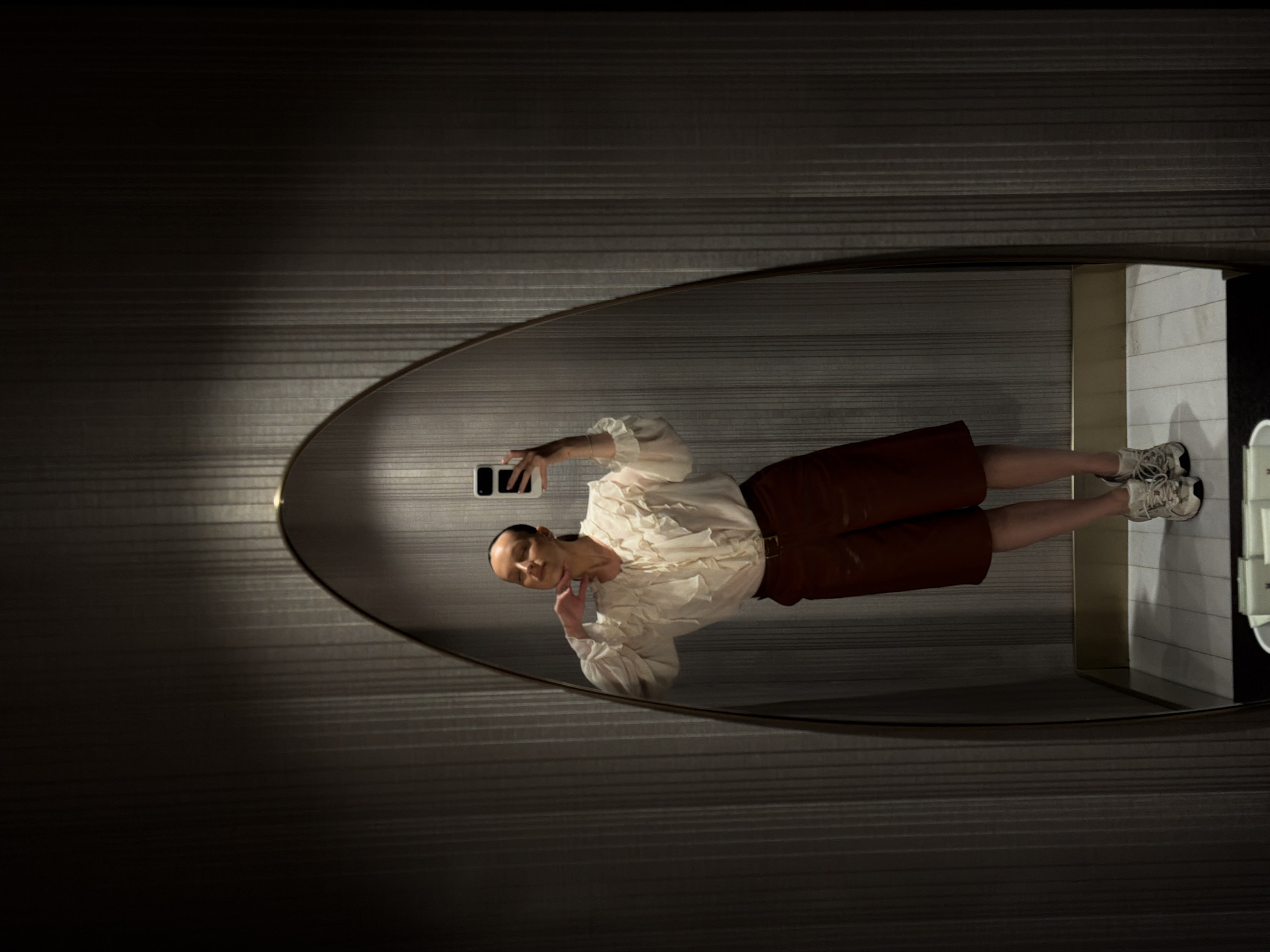 A woman with her hair pulled back stands in front of a full-length mirror, taking a selfie with her smartphone. She is wearing a cream-colored blouse with ruffles and balloon sleeves, a dark red skirt, and white sneakers. She has her right hand on her chin, tilting her head slightly upwards. The mirror is framed by a dark wall with vertical ribbed texture. The floor is tiled in a light color. The setting appears to be a dimly lit restroom, possibly in a hotel or restaurant in Budva, Montenegro.