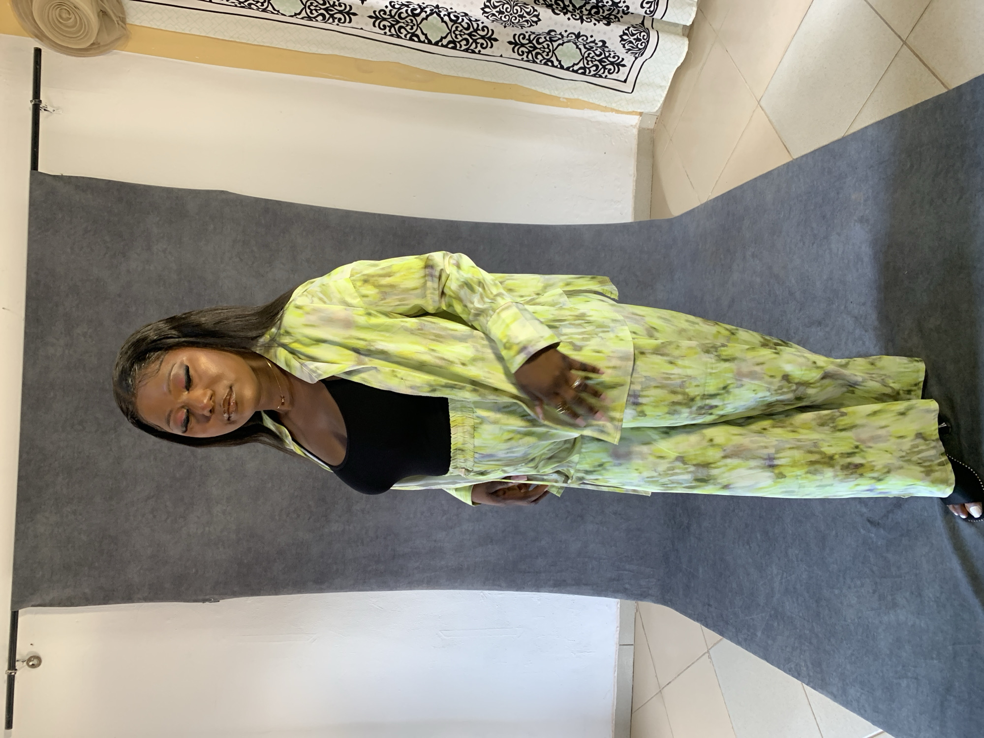 A young Black woman stands with her eyes closed, wearing a black tank top underneath an oversized, light green tie-dye button-down shirt. She also wears matching wide-leg pants. The woman is standing on a gray backdrop, which appears to be in a studio setting. To the right of the backdrop, a portion of a white wall is visible, along with a beige and black patterned curtain hanging. The floor is tiled with light beige square tiles. The setting is consistent with a photography studio located in Jos, Nigeria.