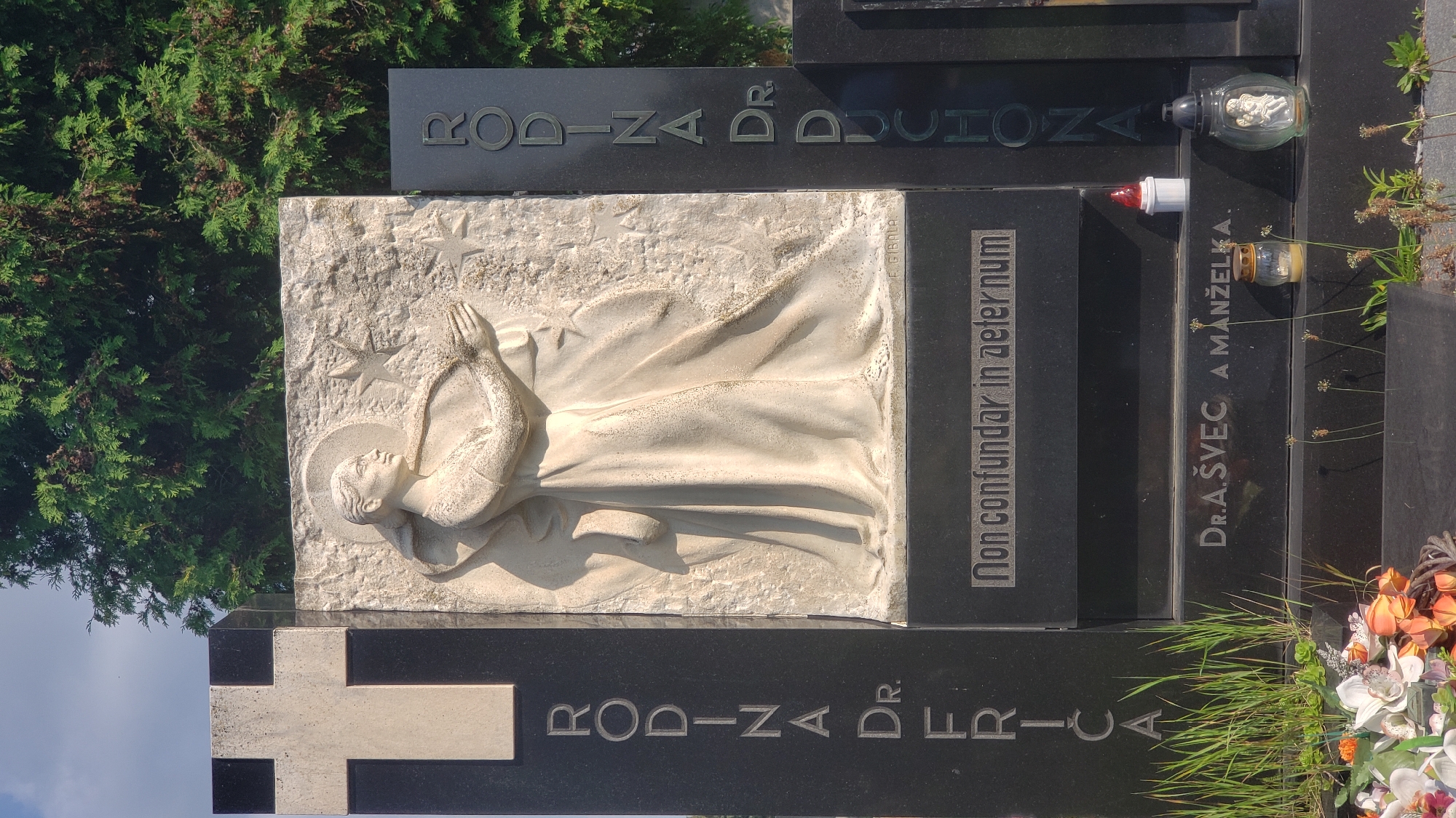 The image shows a gravestone in a cemetery, likely in Prešov, Slovakia, given the names and text. The central element is a large, light-colored relief carved into a stone slab, depicting a woman in flowing robes reaching towards stars. To the left of the relief is a tall, black granite obelisk with a white cross inset at the top and the vertically inscribed text "RODINA DR. FRICA". To the right of the relief is another black granite slab with the vertically inscribed text "RODINA DR. DUCHONA". Below the relief, a black granite plaque is inscribed with "Non confundar in aeter num". Further down, another inscription reads "DR. A. ŠVEC A MANŽELKA". The scene is set outdoors with a tree providing shade from the sky. Various grave decorations, including a small candle, a lantern with an angel figurine, and artificial flowers, are visible at the base of the gravestone. The lighting suggests it is daytime, with shadows indicating the sun's position.