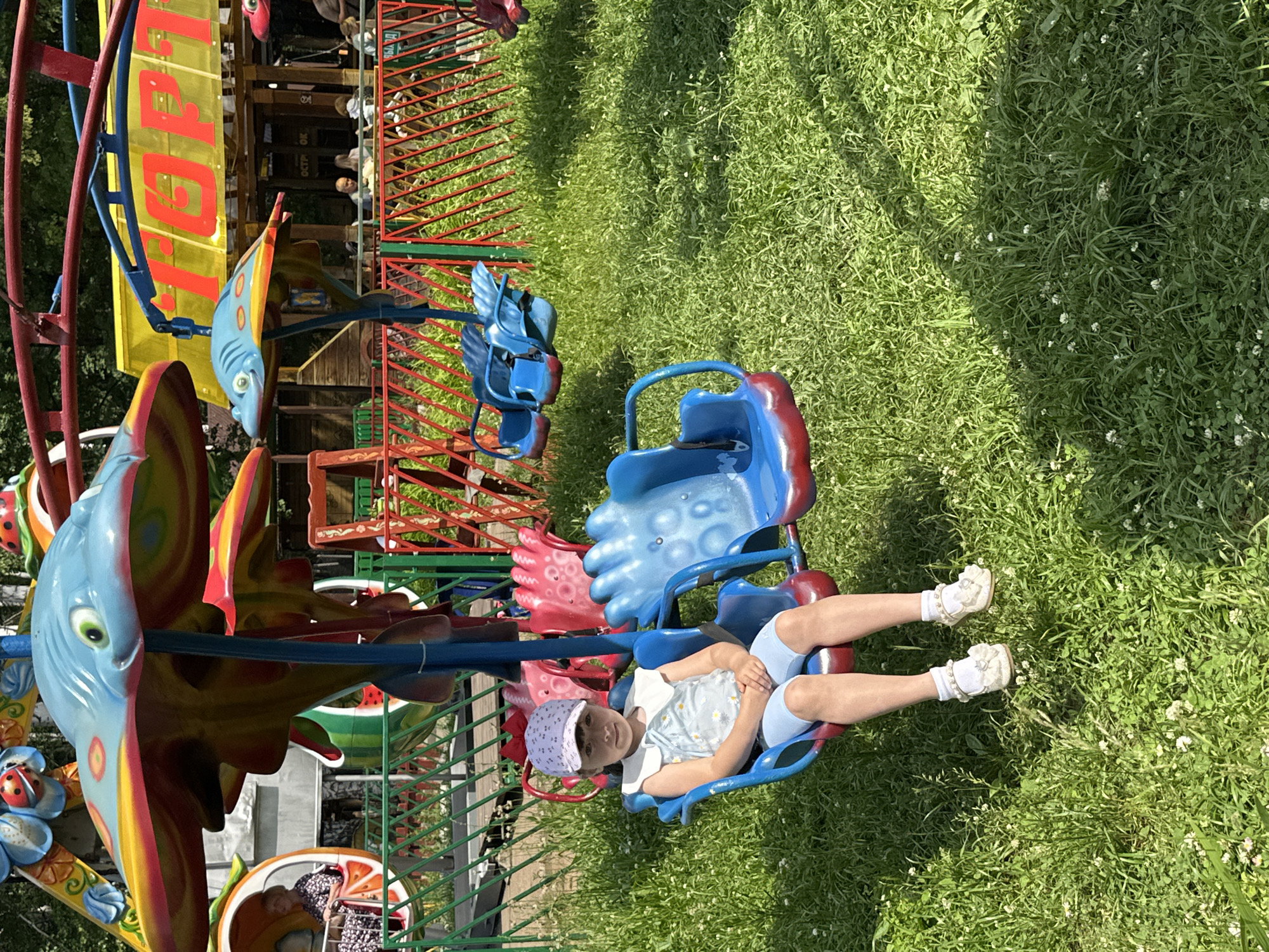 In the city of Chaikovsky, Russia, on a sunny day, a young girl with dark hair and a light-colored cap sits alone in a blue amusement park ride seat. She is wearing a light blue sleeveless shirt with a floral pattern and white collar, and light blue shorts. Her white socks are adorned with small bows and pearl-like beads, and she wears white dress shoes. The ride itself features colorful, whimsical animal-shaped elements, with a blue dolphin-like figure prominently displayed overhead. The girl is positioned in the foreground, with other empty seats of the ride visible around her. In the background, a yellow awning with red lettering, possibly a sign, is partially visible above a structure that appears to be part of the amusement park's amenities. A green metal fence runs through the scene, separating the ride area from the surrounding grassy parkland. The ground is covered in lush green grass dotted with small white flowers. Other people are visible in the distance, suggesting this is a public recreational area.