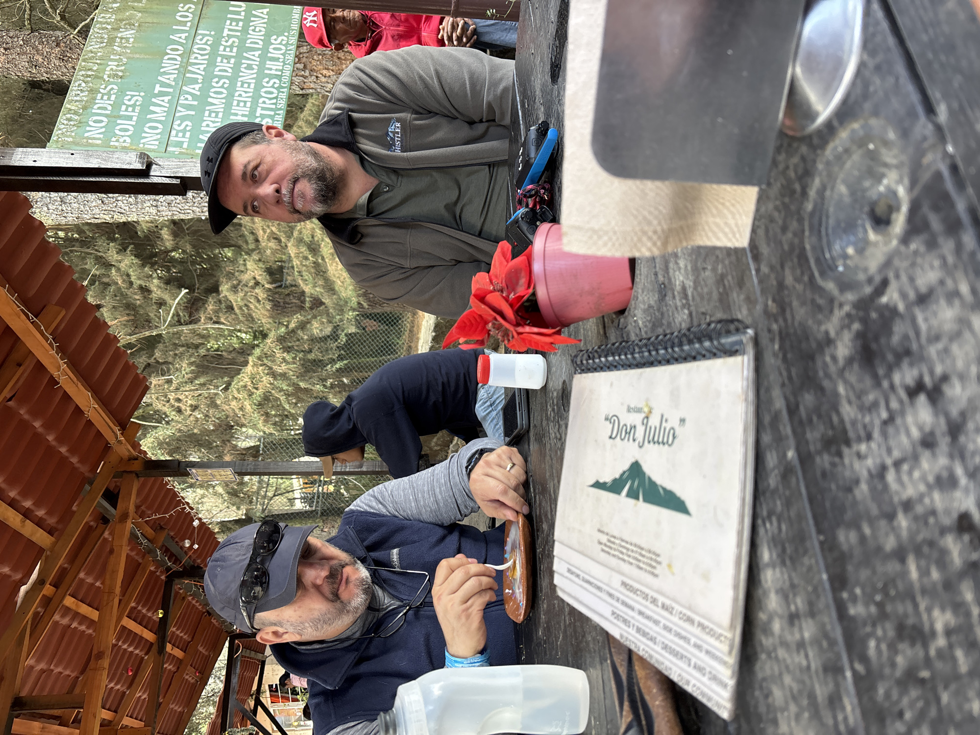 The image captures an outdoor dining scene. Two men are prominently featured, one wearing a blue jacket and a baseball cap with sunglasses resting on the brim. He is engaged in eating something from a small plate with a spoon. The other man, wearing a gray jacket and black baseball cap, is looking directly at the camera. A third person, wearing a red jacket and hat, is partially visible in the background.
The setting appears to be a restaurant named "Don Julio," as indicated by a menu on the table. The table itself is wooden and has a red potted plant on it. A sign with Spanish text is also visible, appearing to be a conservation message. The environment includes trees, a glimpse of a building with a reddish roof, and what appears to be the edge of a forest.
Based on the text on the sign ("NO DESTRUYENDO L BOLES!") and further text, and the presence of the restaurant "Don Julio," this is in San José Teacalco, Mexico.