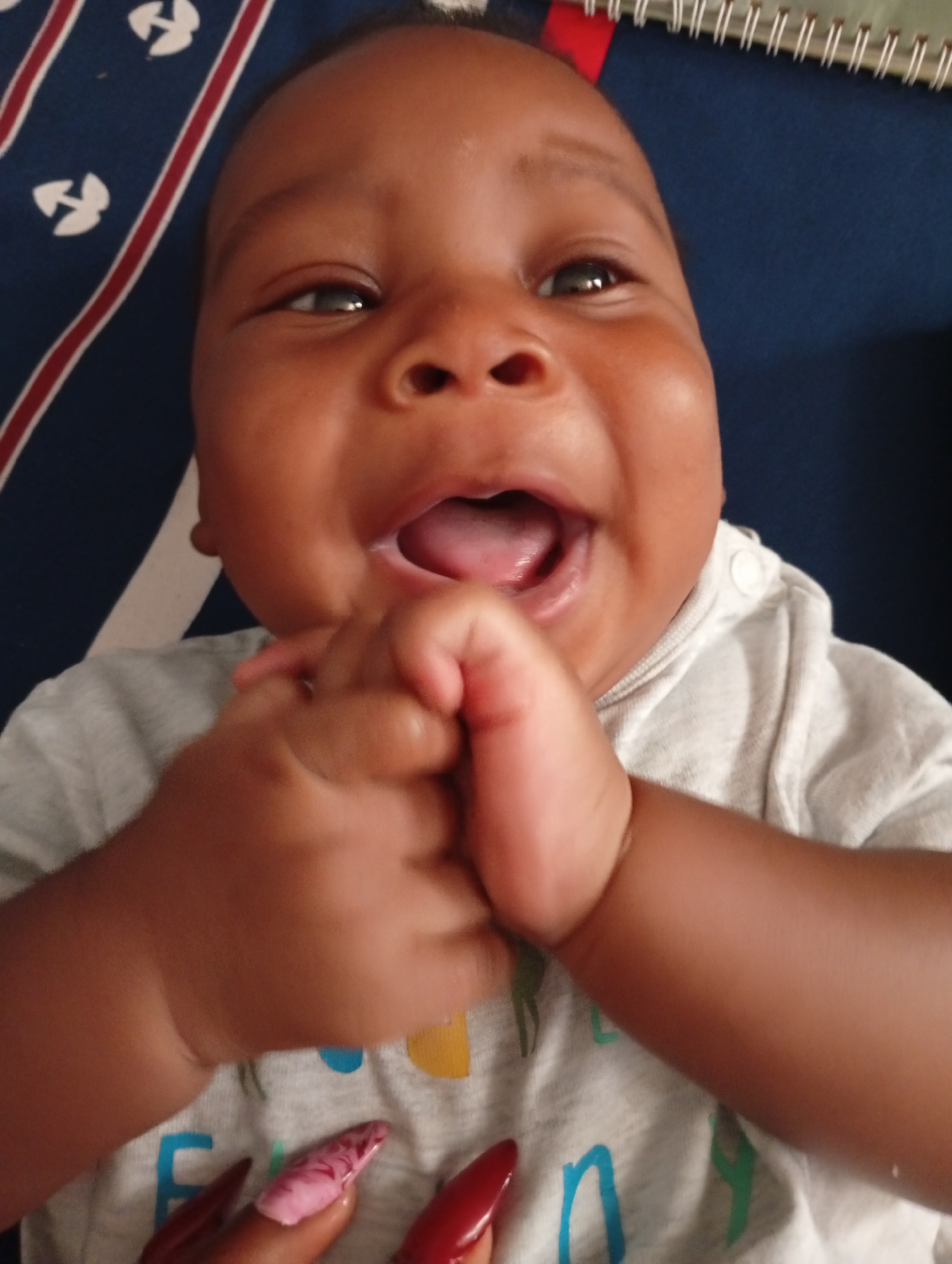 A baby with dark skin, wearing a light gray shirt, is lying on a blue surface patterned with white shapes and red stripes. The baby is smiling with their mouth open and hands clenched together. A hand with long, red and pink manicured nails is visible in the lower left corner. A notebook is in the upper right corner. It is not possible to determine the location is Sabon Tasha, Nigeria from the image.