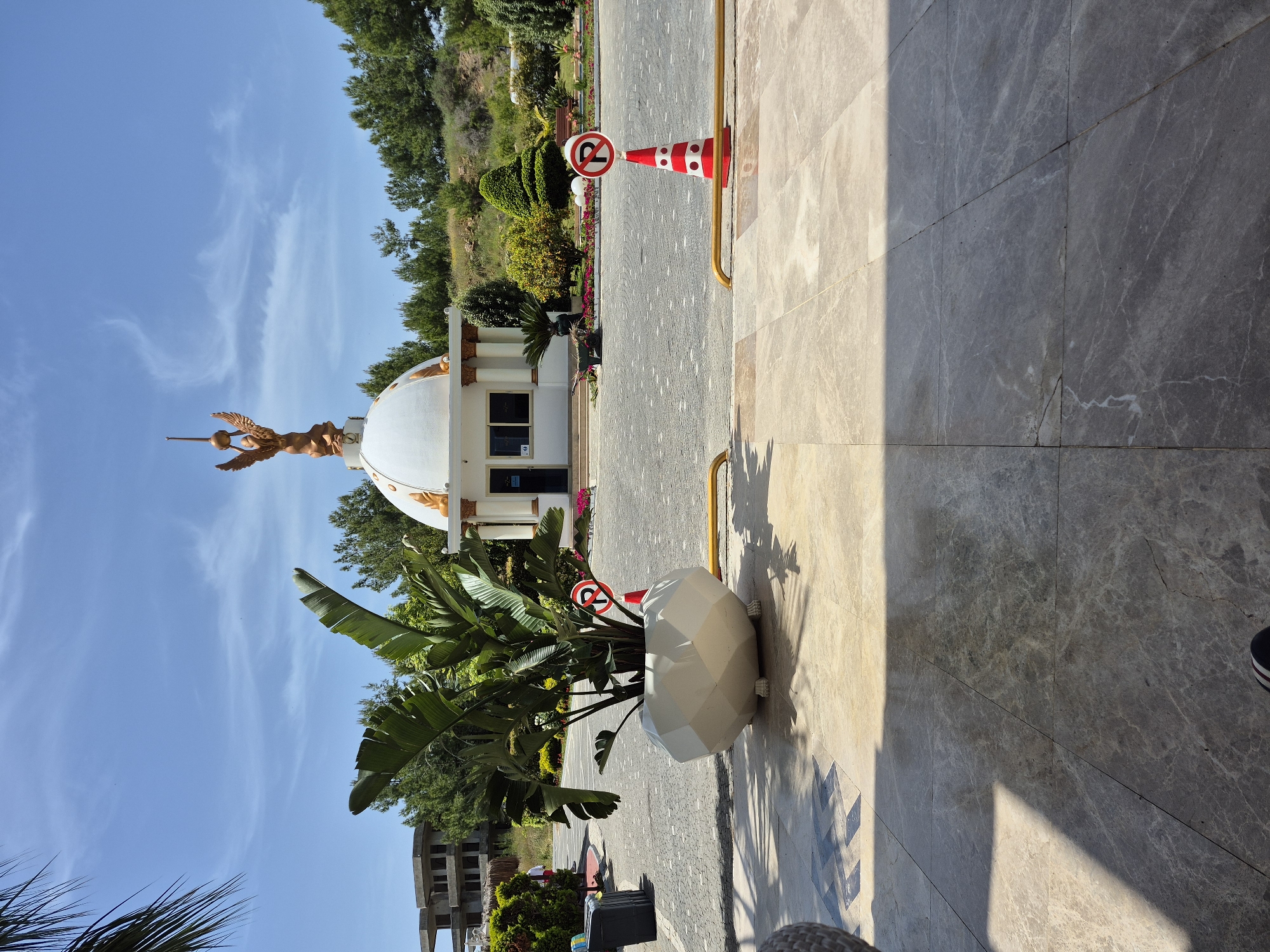 The image depicts an outdoor scene in Alanya, Turkey, featuring a white, dome-topped building with a gold-colored statue of an angel on top. The statue holds a sphere above its head, supported by a spear-like object. The building is adjacent to a paved area, likely a pedestrian walkway or road.
A "no parking" sign is visible twice in the image. One sign is attached to a red and white striped traffic cone, while the other is next to a white, geometric-shaped planter containing lush, green banana plants.
The background includes trees, shrubs, and other vegetation, suggesting a park-like or landscaped setting. The sky is blue with scattered clouds.
The image appears to be taken from the perspective of someone standing on or near a tiled or marble-paved surface, with some shadows cast on the surface.