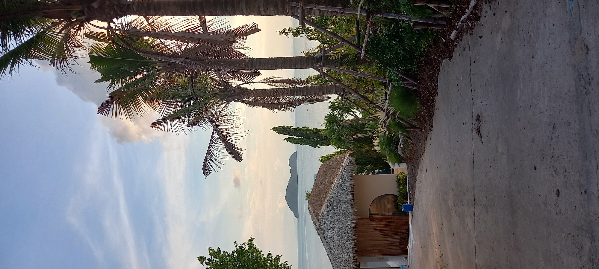 The image shows a coastal scene in Baan Wok Tum, Thailand, featuring palm trees, a building with a thatched roof, and the sea. The sky is visible with clouds, indicating either sunrise or sunset. A concrete path or road runs along the right side of the frame. The building appears to be a structure near the beach with an arched doorway. In the distance, a landform, possibly an island or peninsula, is visible in the sea. The vegetation along the path includes various plants and shrubs.
