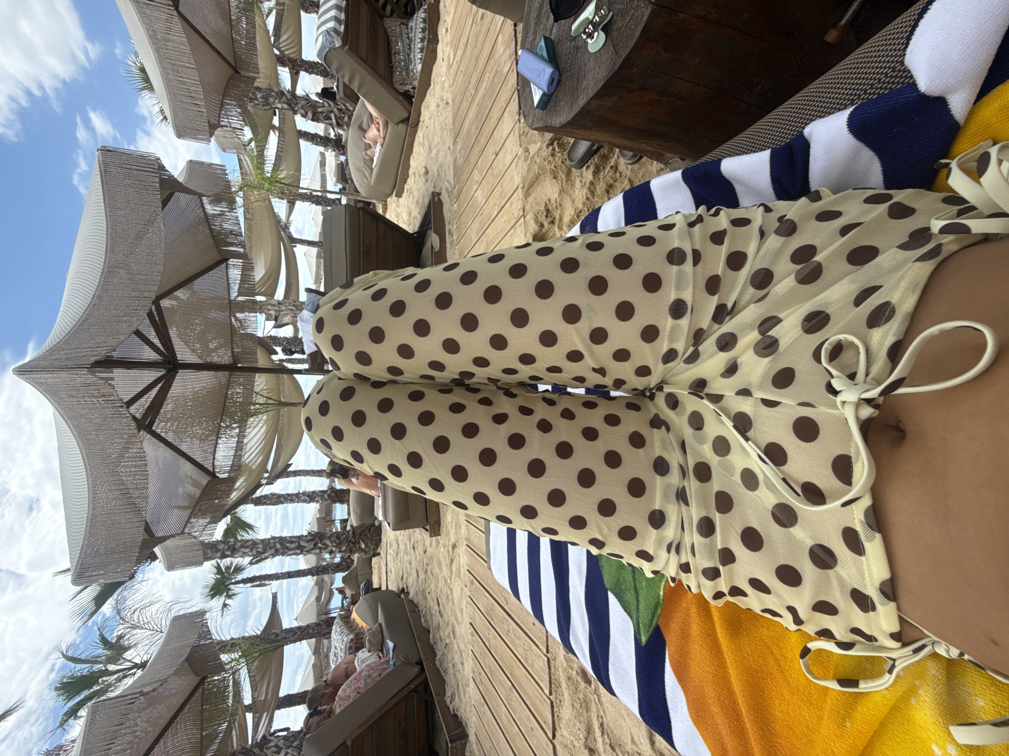The image shows a person's legs, clad in sheer, polka-dot pants, resting on a striped beach towel. The towel is placed on a sun lounger at Ofrynio Beach in Greece. In the background, several other sun loungers and palm trees are visible, along with beach umbrellas. The sand is a light brown color, and the sky is partly cloudy with a blue hue. A wooden table with a blue phone and a pair of sunglasses rests to the right of the person. The overall scene depicts a relaxed day at the beach.