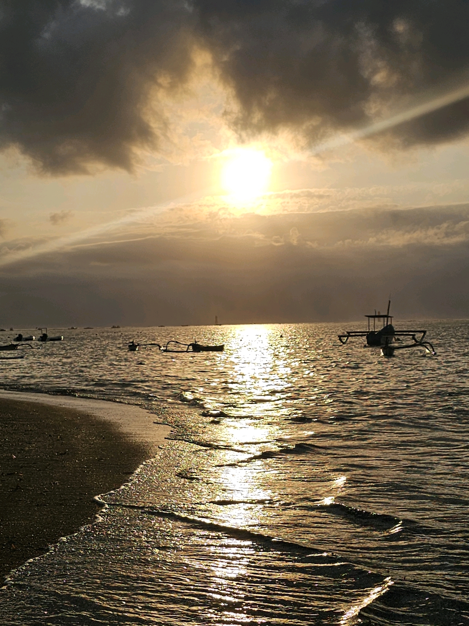 The image depicts a tranquil scene at Sanur Beach in Denpasar, Indonesia, during sunset. The sky is a dramatic canvas of dark, heavy clouds tinged with the warm hues of the setting sun, which is a bright, diffused orb peeking through the clouds, casting a golden glow on the water. The sun's rays are visible, streaking across the sky and reflecting brilliantly on the gentle ripples of the ocean. Several traditional Balinese fishing boats, known as jukungs, are scattered across the water. One jukung, with its characteristic outriggers, is prominently featured in the mid-right of the frame, bobbing gently. Another jukung is visible further to the left, and more distant boats are suggested by their silhouettes against the shimmering sea. The shoreline in the foreground is composed of dark sand, with the soft lapping of waves creating a gentle foam line. The overall atmosphere is one of peaceful serenity, capturing the essence of a typical evening on this popular Indonesian beach.