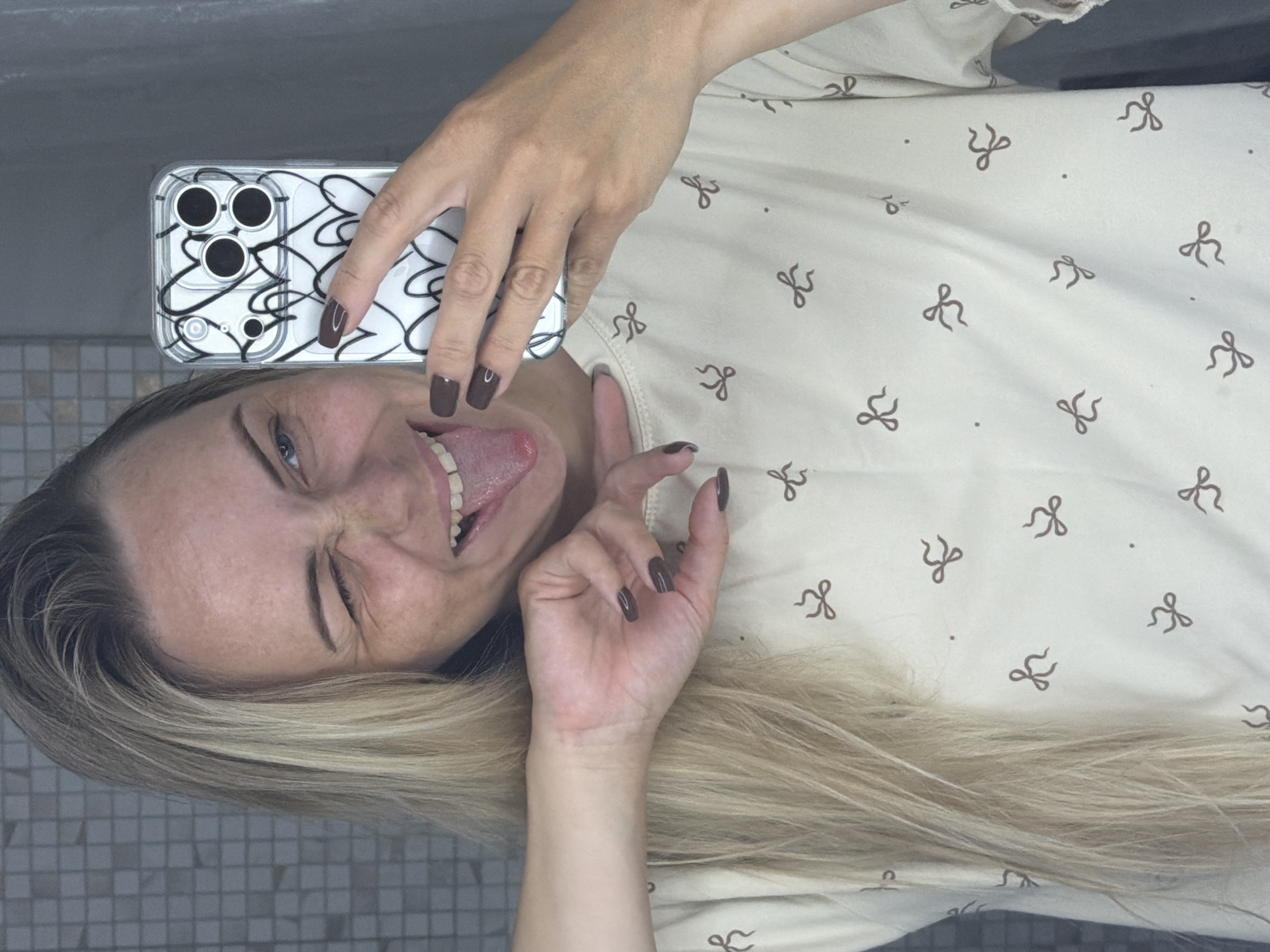 This is a selfie taken by a person in Votkinsk, Russia. The person is holding a smartphone with a clear case featuring a black scribbled design. They are winking and sticking their tongue out, suggesting a playful or humorous mood. The person has long blonde hair and is wearing a light-colored top with a repeating pattern of small brown bows. The background appears to be a tiled surface, possibly a bathroom or kitchen. The lighting suggests it is daytime.