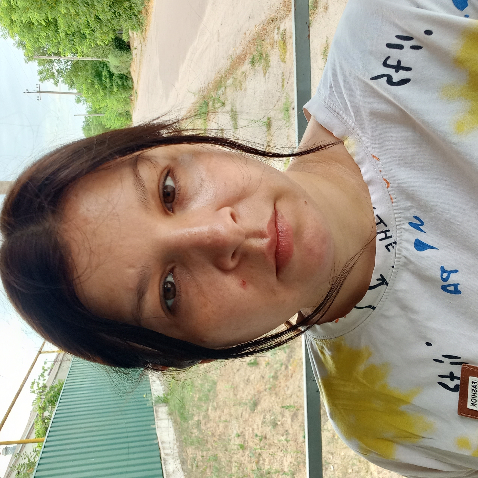 In a medium close-up selfie, a woman with dark hair and brown eyes looks directly at the camera. She is wearing a white t-shirt with yellow and blue splotches, and some text printed on it. The background is blurred, but appears to be an outdoor setting with a dusty road and green trees. There are utility poles visible in the distance. The photo was taken in Klyntsi, Ukraine.