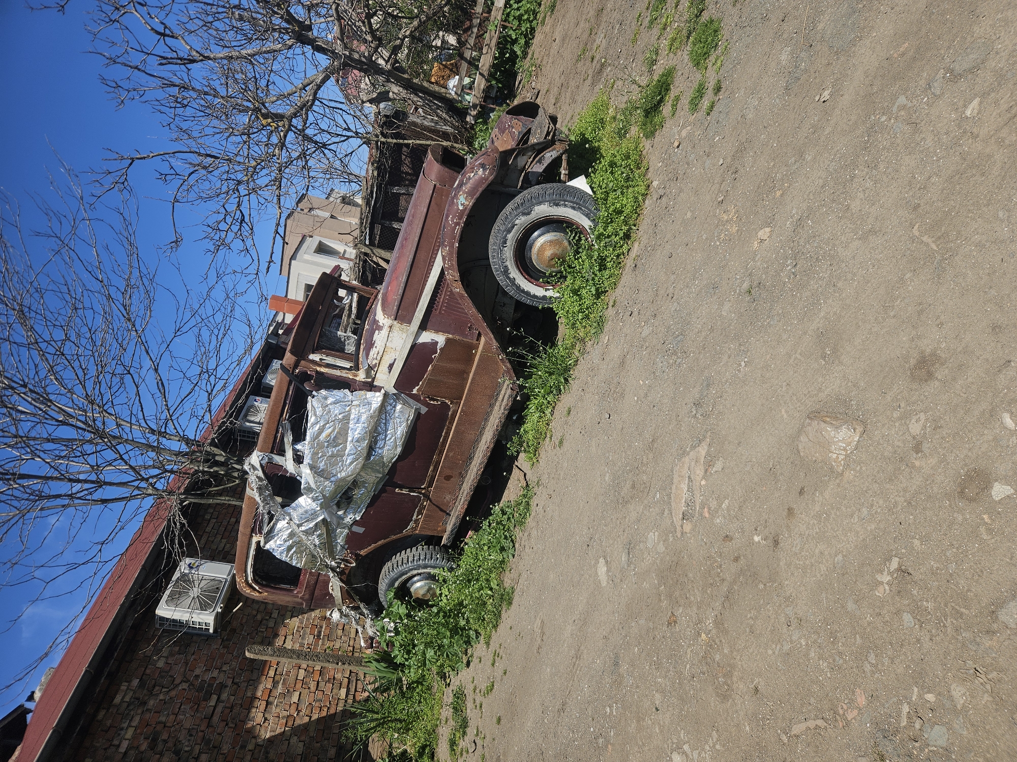 The image shows a heavily rusted and dilapidated vintage vehicle, possibly a truck, parked beside a brick wall and an unpaved road in Burgas, Bulgaria. The vehicle is heavily overgrown with greenery, and part of its interior is covered with shiny, possibly metallic, material. The sky is clear and blue, visible through the bare branches of a tree. A building is also visible in the background. A modern air conditioning unit is attached to the brick wall.