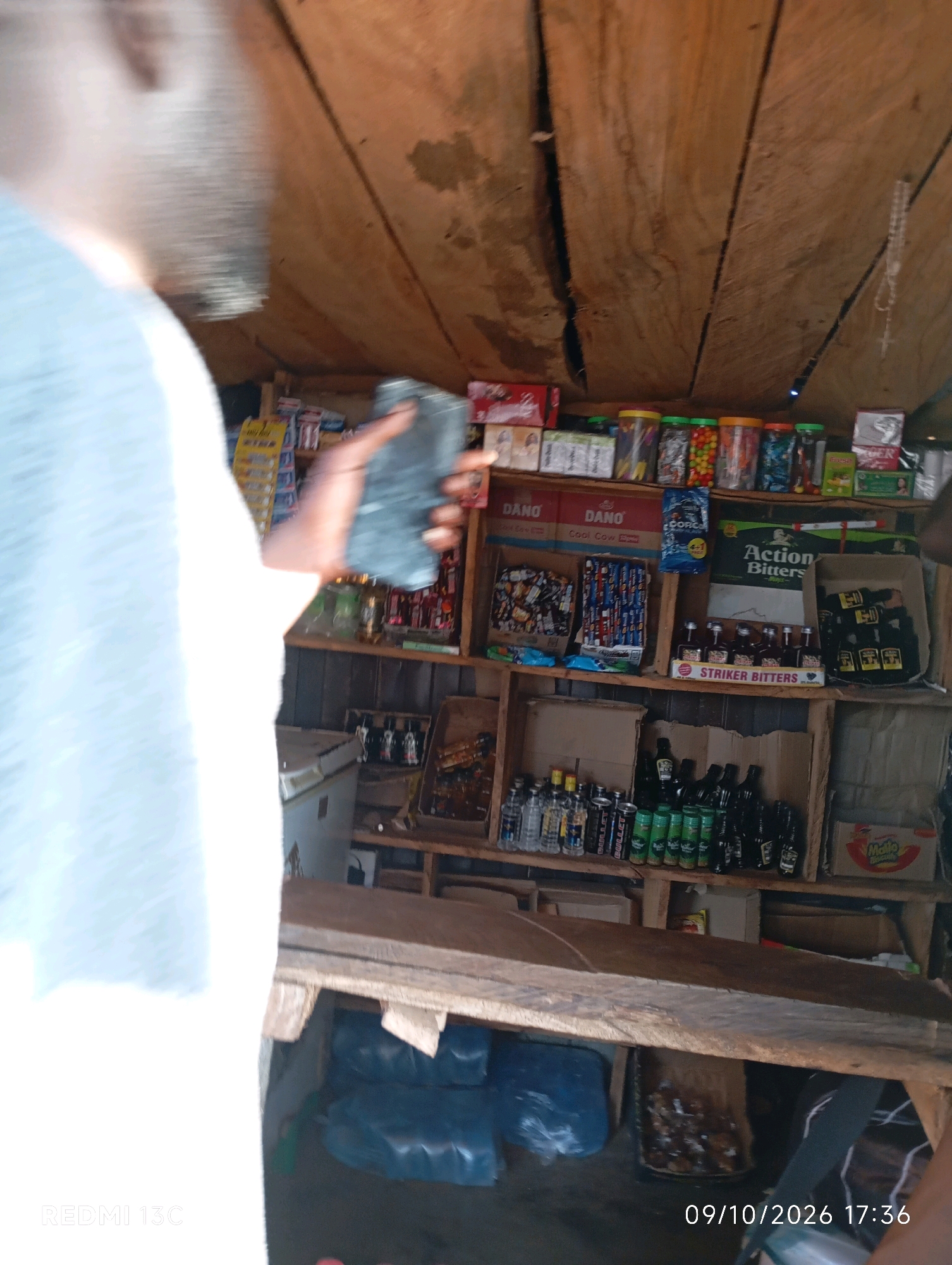 The image shows a dimly lit stall with wooden shelves displaying various items, likely in Shendam, Nigeria. A person, partially visible on the left, is holding a dark, cylindrical object, possibly a bottle or a can, in their right hand. The shelves are stocked with a mix of products, including what appear to be drinks, snacks, and small packaged goods. Several bottles of what looks like alcohol are visible, some labeled "Striker Bitters." In the background, there are also larger plastic-wrapped items and cardboard boxes. The overall environment suggests a small convenience store or a roadside vendor stall. The lighting is poor, and the image quality is grainy. A date and time stamp in the bottom right corner reads "09/10/2026 17:36".