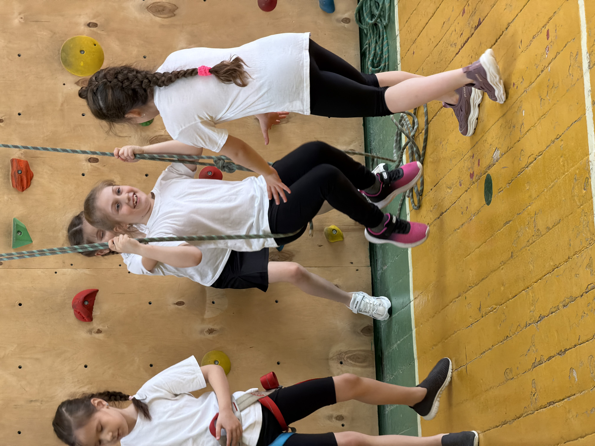 The image shows four girls on a climbing wall. The wall has climbing holds of various colors. The girls are wearing white t-shirts and dark-colored athletic bottoms. Some are using a rope. The background consists of the climbing wall and what appears to be a gymnasium floor. The location and city of Izhevsk, Russia, are not identifiable from the image.