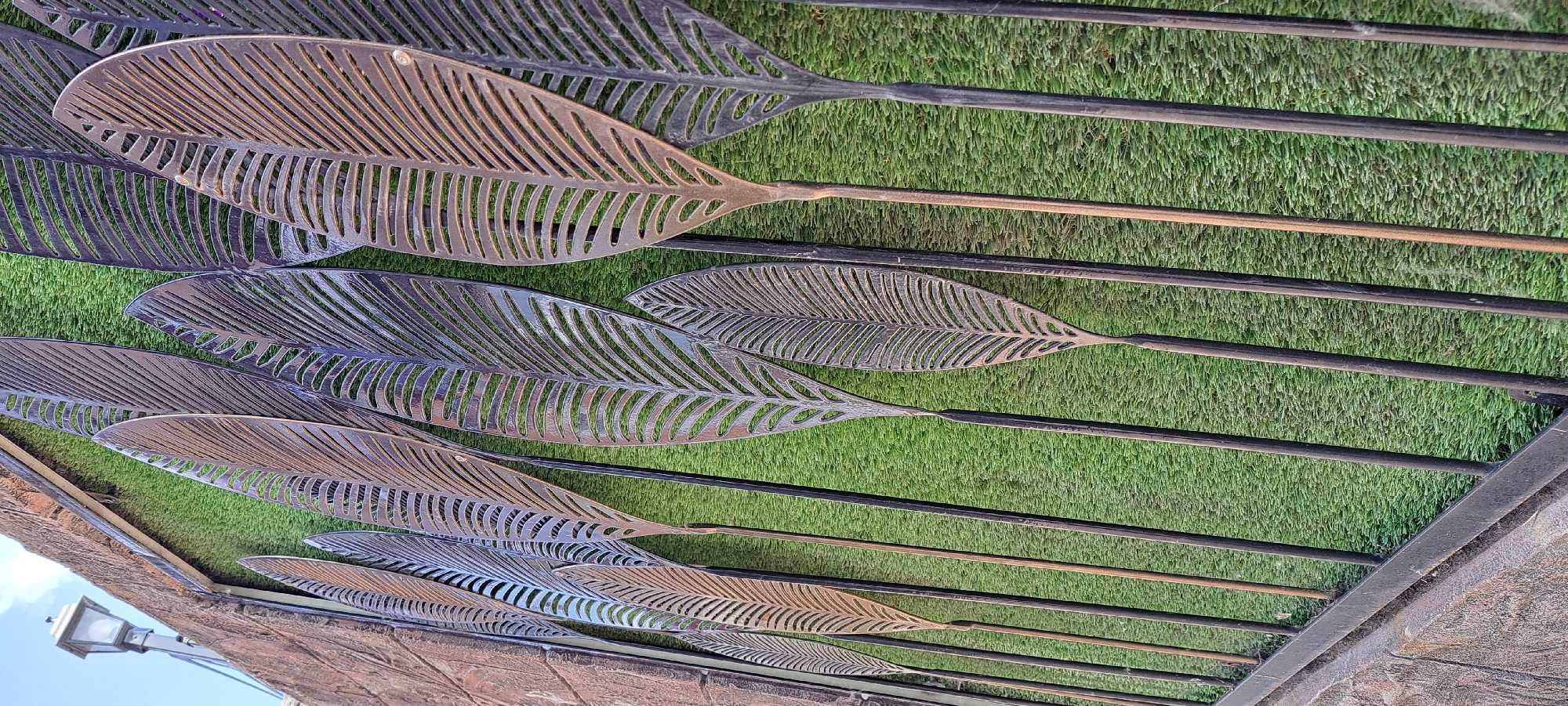 The image shows an outdoor decorative wall art installation in Yessentuki, Russia. The art consists of multiple large, stylized metal leaf shapes with intricate cut-out patterns, resembling palm or fern fronds. These metal leaves are mounted vertically on dark, slender metal rods, creating a layered effect against a backdrop of artificial green turf. The turf is textured and appears to mimic natural grass. To the left, a rough-textured, reddish-brown stone wall is visible, with a lamp post attached to it at the top left corner. The overall impression is one of a modern and artistic exterior decoration, possibly adorning a building or a garden area. There are no people or animals present in the image.
