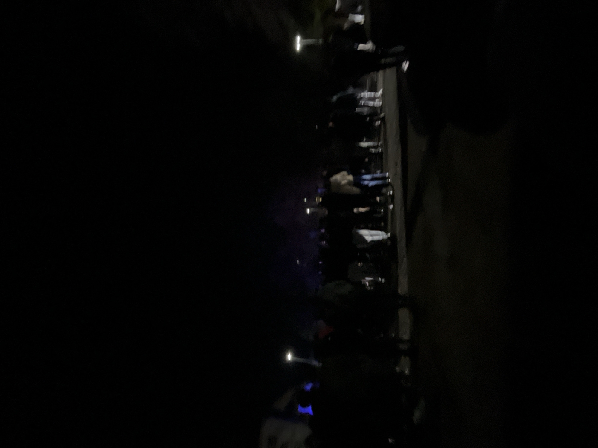 The image depicts a dimly lit street scene at night. A crowd of people is gathered on the right side of the frame, illuminated by scattered streetlights. The background is dark, obscuring the details of the buildings or environment. The location in the city of Burgas, Bulgaria cannot be determined based on the provided image.