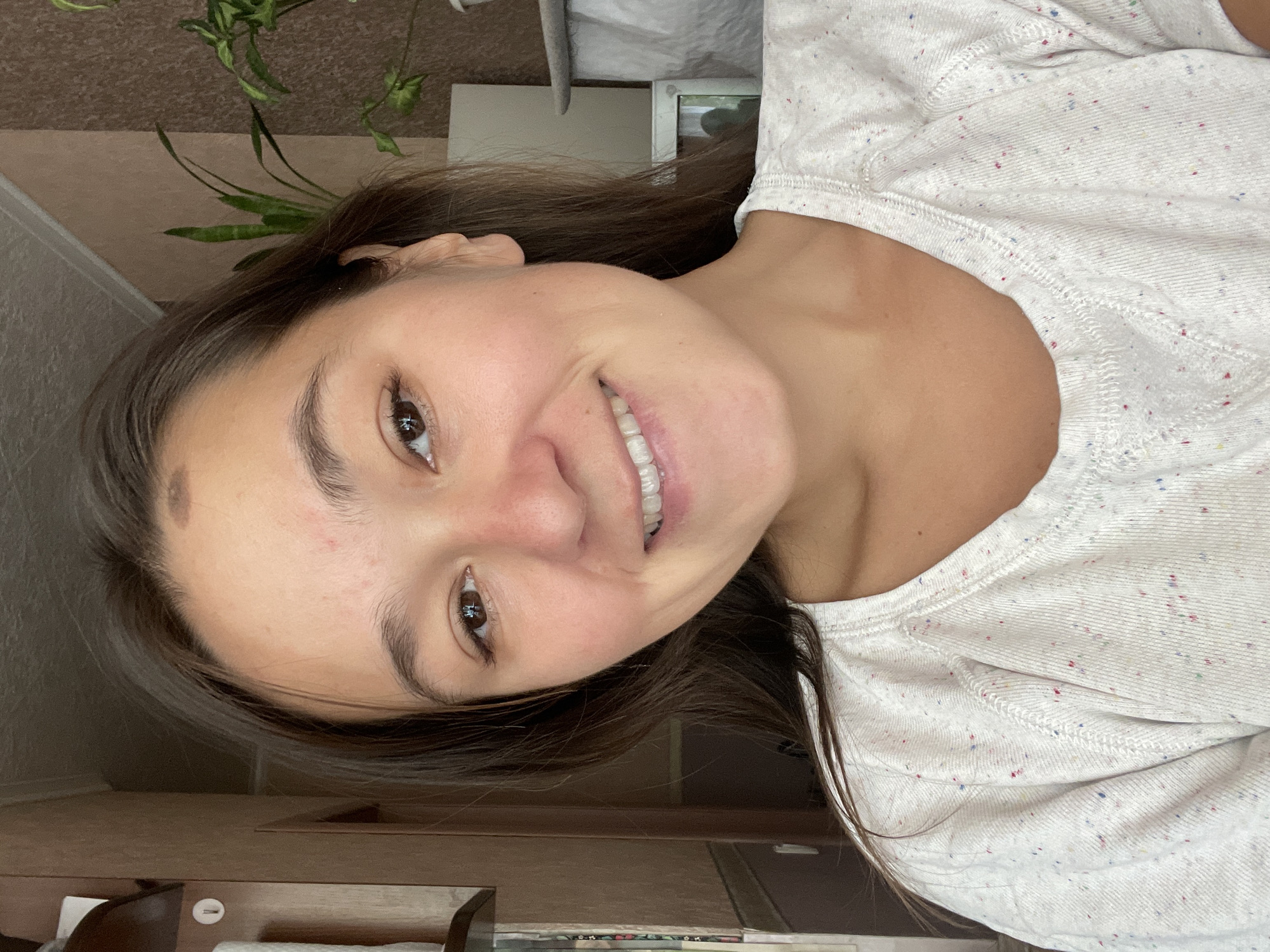 The image shows a young woman indoors, likely in a residential setting in Zheleznogorsk, Russia. She is positioned in the foreground, facing the camera and smiling. The woman has brown hair and is wearing a white t-shirt with colorful flecks. The background reveals elements of a room, including a wall with patterned wallpaper, a piece of furniture that appears to be a shelf or cabinet, and some potted plants. The lighting suggests it is daytime.