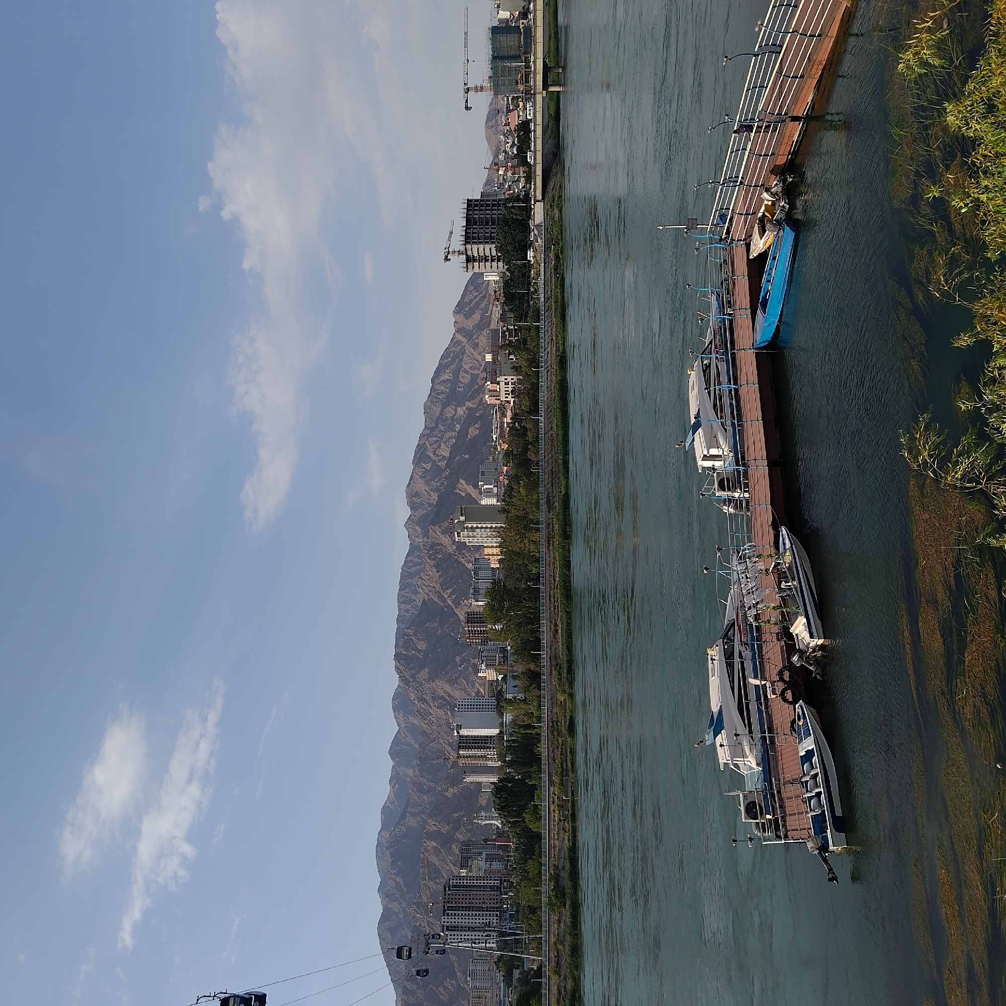 The image depicts a panoramic view of the city of Khujand, Tajikistan, with a focus on its waterfront and skyline. In the foreground, a wide, greenish river flows, with a wooden pier extending into it. Several boats, including speedboats and smaller dinghies, are moored to the pier. The pier itself has railings and lights, suggesting it's a well-maintained recreational area.
In the mid-ground, a treelined embankment separates the river from the city. The city skyline is dominated by modern, multi-story buildings, some appearing to be under construction with cranes visible. A prominent cable car system with multiple cabins is seen ascending towards the mountains in the background.
The background features a rugged, arid mountain range that frames the city. The sky above is a clear blue with scattered white clouds. The overall scene suggests a dynamic urban environment situated against a dramatic natural landscape, with elements of recreation and development coexisting.