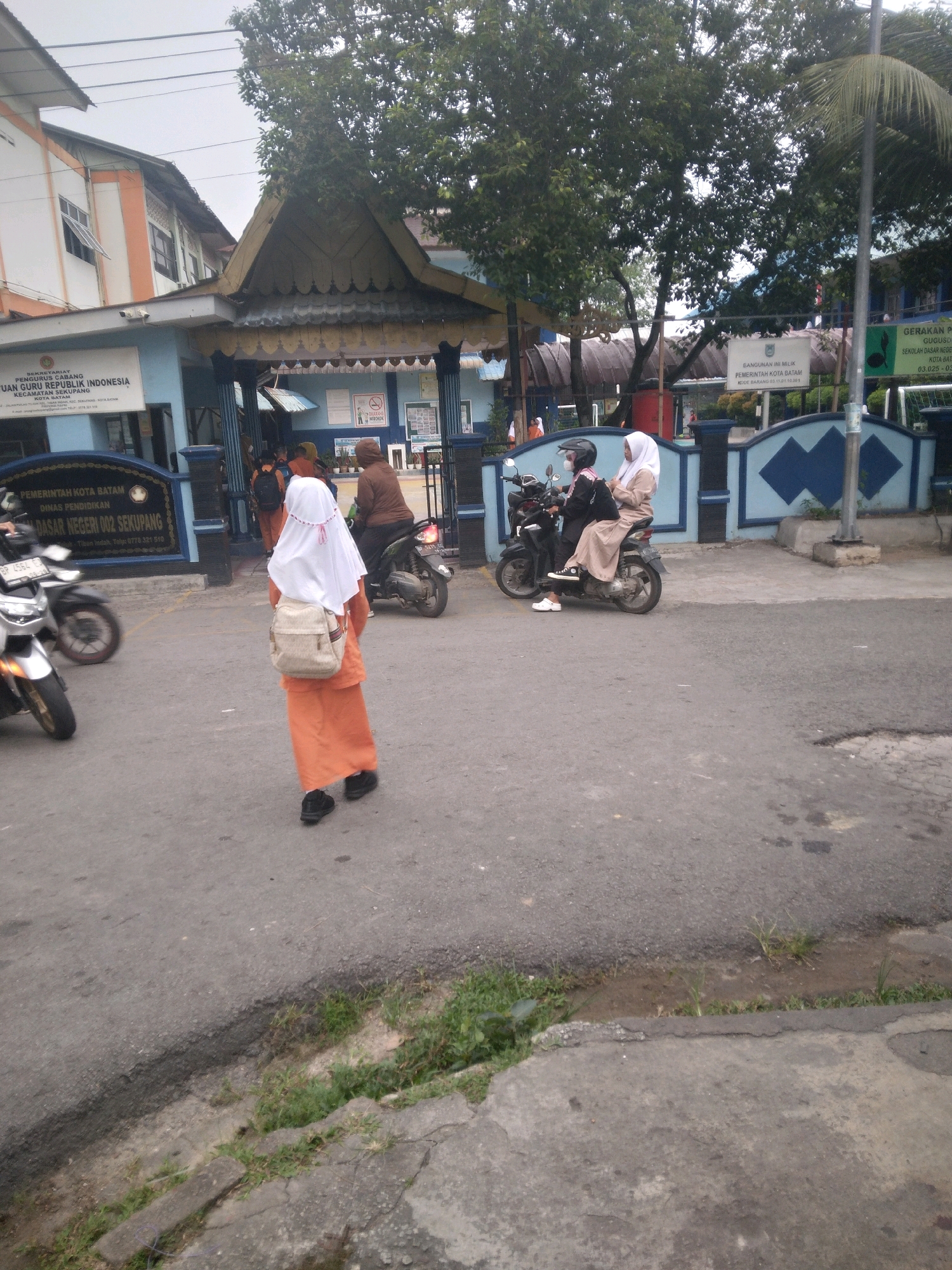 The image depicts the entrance to a school in Batam, Indonesia. A young student, wearing an orange skirt, white hijab, and carrying a beige backpack, walks away from the camera on a paved road. Behind her, two individuals are riding a motorcycle, the passenger wearing a white hijab and the driver a black helmet and mask. Further in the background, other students and adults are visible near the school entrance, with motorcycles parked outside. The school building has a blue facade with signs that appear to be in Indonesian. The overall environment suggests a typical school day scene in a humid, tropical climate, with trees and a cloudy sky overhead.