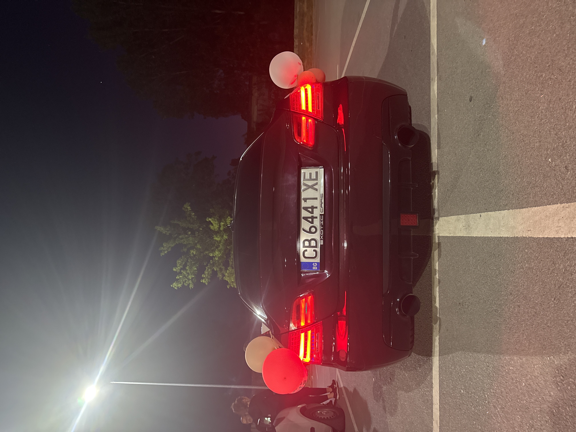 The image shows the rear of a dark-colored car parked on an asphalt surface with white parking lines. The car has a Bulgarian license plate reading "CB 6441 XE." There are two balloons attached to the rear of the car, one red and one white. The car's taillights are illuminated. In the background, there are trees and streetlights visible. In the lower part of the image, there are some people and a part of a car. The environment appears to be urban. The image was likely taken at night. The location of the city is unknown, Unknown.