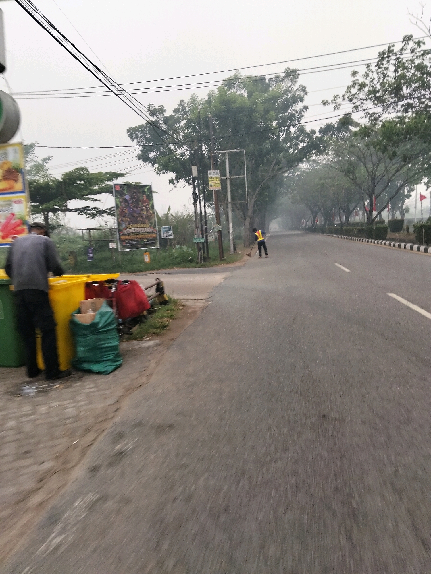 The image depicts a street scene in Teluk Kapuas, Indonesia, characterized by a moderately hazy atmosphere and overcast sky. On the left side of the road, a person wearing a grey jacket and black pants stands near a collection of waste bins: a large green bin, a yellow bin, and a red bag and a green bag of trash. Further down the sidewalk, a person in a yellow safety vest is sweeping the side of the road with a broom. The road itself is paved and appears to be a two-lane street with white lane markers. The right side of the road is bordered by a curb with black and white stripes, followed by a row of manicured bushes and trees. Several red flags are visible on poles along the roadside, suggesting a potential event or commemoration. In the background, trees line the street, their branches creating a canopy over the road, and the haze contributes to a softened, diffused lighting condition. A large poster with text and images is affixed to a pole on the left side of the scene, near the waste bins.