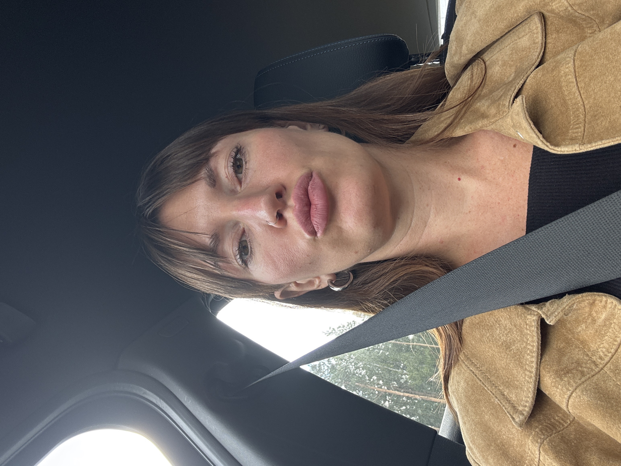 The image shows a woman in the driver's seat of a car, with a seatbelt fastened. She has medium-length brown hair, bangs, and is wearing a light brown suede jacket over a black top. Her mouth is puckered, and she is looking directly at the camera. The background shows trees and a cloudy sky, suggesting an outdoor setting. The car interior is visible, including the dashboard and part of the passenger seat. There are no other people or animals in the image. The location is identified as Pushkino, Russia.