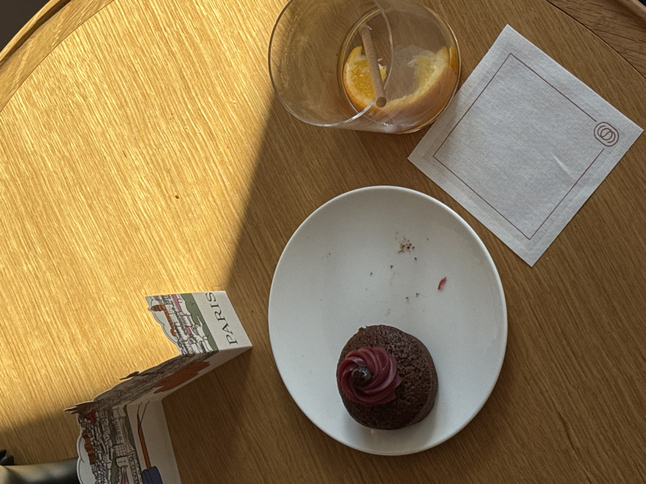 An overhead view displays a setting on a light-colored wooden table, possibly a cafe or restaurant, in an unknown city. The table surface exhibits a distinct wood grain and is partially illuminated by a strong, angled light source from the upper right, casting a shadow across the upper left quadrant.
In the center-left, a round white ceramic plate holds a dark brown cupcake or muffin topped with a swirl of reddish-purple frosting and a single dark berry. The plate shows evidence of prior consumption, with crumbs and a reddish-purple smear present.
To the right of the plate, a clear glass contains a light-colored liquid. Inside the glass are a large orange peel and a single cinnamon stick. The liquid level in the glass is below full.
Below the glass, a white square paper napkin is positioned, featuring a thin reddish-brown border and a small, stylized circular logo (two interlocking circles) in its bottom right corner.
Behind the plate, partially in the shadow, stands a small, folded white card or menu item. One visible panel displays a line drawing of buildings and the word "PARIS" printed on its base. No individuals are visible in the scene.
