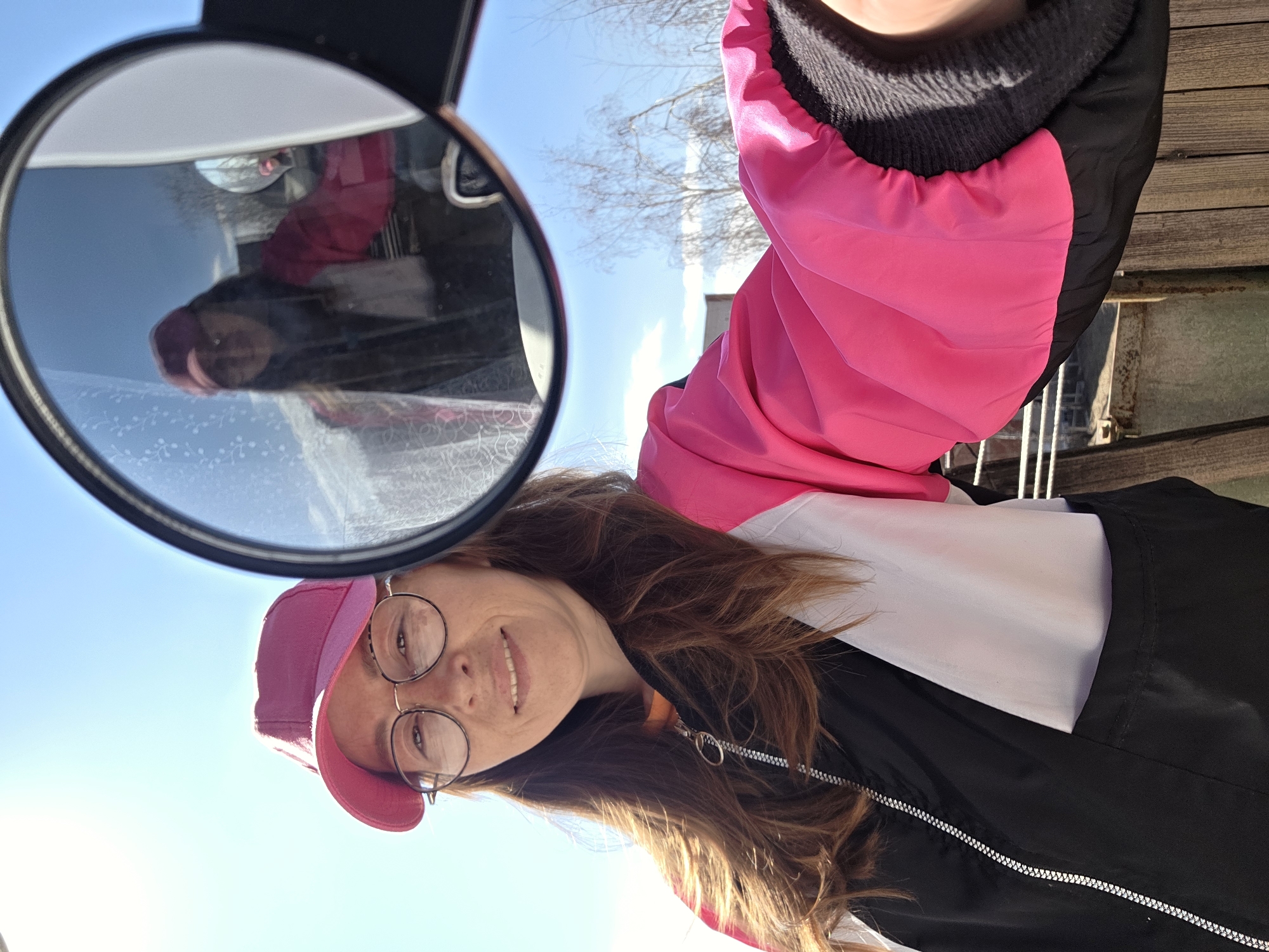 This outdoor portrait photograph, originally rotated 90 degrees clockwise, captures a woman with long, reddish-brown hair, wearing a pink baseball cap and round, wire-rimmed glasses. She displays a gentle, pleasant smile. She is dressed in a layered outfit consisting of a black outer jacket with a distinct white section across the chest, worn over a bright pink hooded garment with a visible black knitted collar.
A circular mirror, positioned in the upper right quadrant of the image (when viewed upright), reflects the woman. In the reflection, she is also seen wearing the pink cap and a pink top, maintaining a faint smile.
The background features a clear, bright blue sky with sparse, wispy white clouds. Bare tree branches are visible against the sky, suggesting the scene takes place in late winter or early spring. To the woman's right, wooden planks, likely part of a deck or boardwalk, and some metal railing are visible, indicating an outdoor setting.
The strong, direct sunlight suggests it is daytime, possibly mid-morning or afternoon. There is no visible text in the image. The location context for this scene is Chaikovsky, Russia.