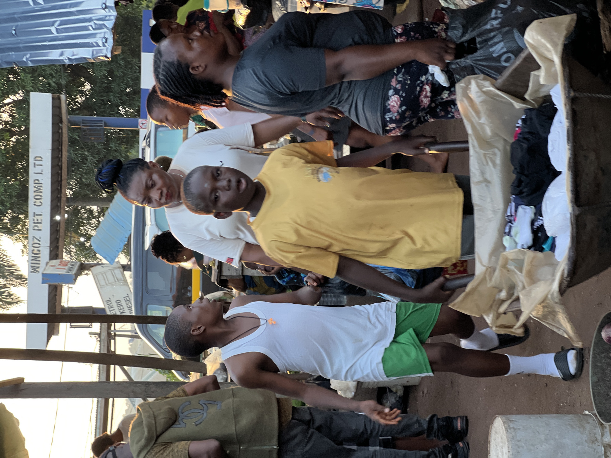 This is a daytime, eye-level, medium-shot photograph taken in the city of Nkpor, Nigeria, near a gas station. In the foreground, a young boy in a yellow t-shirt and green shorts walks with his mouth open, looking towards the camera. To his left, another boy wearing a white tank top and green shorts, with a necklace, also looks to the side. Behind them, a woman in a white t-shirt with dark hair pulled up in a bun gazes forward. To the right of the woman in white, another woman in a dark grey t-shirt and floral leggings is seen from the side, holding a black plastic bag. In the background, a dark-colored van is parked, and a sign for "WINGOZ PET. COMP. LTD" is visible. The environment appears to be a market or roadside area, with various objects and people scattered around. The lighting suggests it is either early morning or late afternoon.