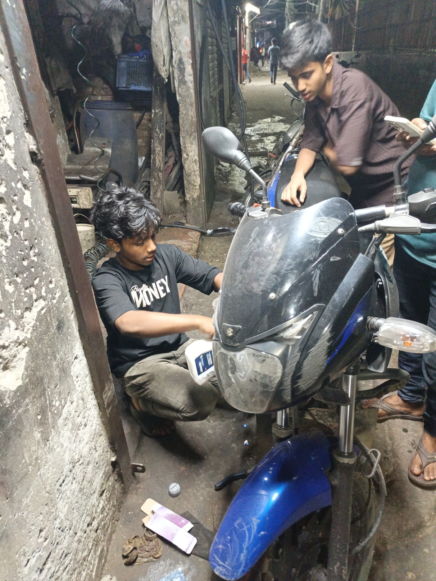 The image depicts a narrow alleyway in Dhaka, Bangladesh, with two young men working on a motorcycle. The alley is cramped, with weathered concrete walls and exposed pipes. One man, wearing a black t-shirt with "Money" printed on it and olive pants, is crouched down, seemingly pouring something from a white container into the motorcycle. The other man, wearing a maroon shirt, stands beside the motorcycle, his hand resting on the fuel tank. A third person wearing a turquoise shirt is partially visible to the right of the frame. The motorcycle is a blue and black Pulsar. Debris and discarded items litter the ground around the motorcycle, including discarded paper, a bottle cap, and a rag. The location appears to be a small mechanic shop within a densely populated urban area. Other people can be seen at the end of the alleyway.