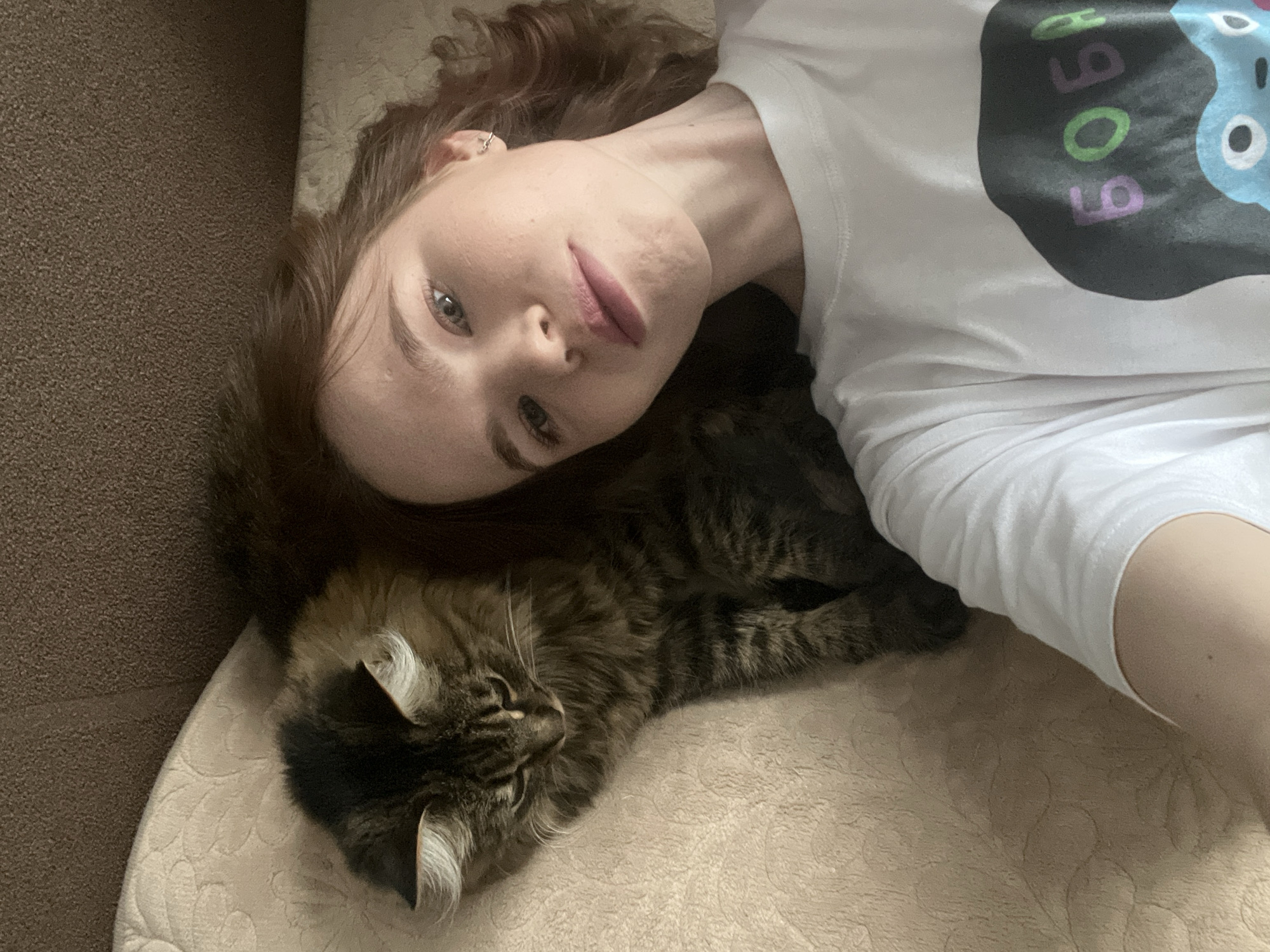 A woman with reddish-brown hair and pale skin is lying on a beige textured surface, likely a bed or sofa. She is wearing a white t-shirt with a colorful, cartoon-like design on the front. Her face is turned towards the camera. A furry, brown tabby cat is lying beside her, its head near her shoulder. The setting appears to be indoors. There is no indication of the city Chaikovsky, Russia.