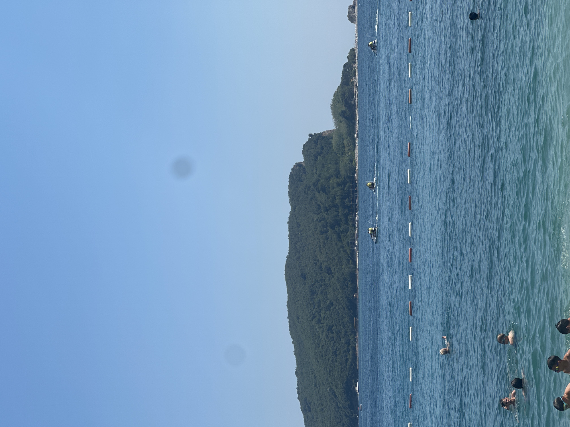 This is a vertical, eye-level, outdoor shot of a beach scene in Budva, Montenegro. The bright blue sky occupies the left half of the frame, with a blurred dark circular spot visible in the sky. The right half of the frame shows the clear blue water of the sea, with a line of orange and white buoys marking a swimming area. The shore is lined with lush green trees and vegetation, with some buildings visible in the distance along the coastline. The time of day appears to be daytime, with bright sunlight.
Several people are visible in the water, swimming and enjoying themselves. Some are partially submerged, while others are fully visible. There are at least two jet skis in the water near the shore, with riders on them. One jet ski appears to be moving at speed, creating a wake. The overall mood of the scene is one of leisure and recreation on a sunny day at the beach. There is no visible text in the image.