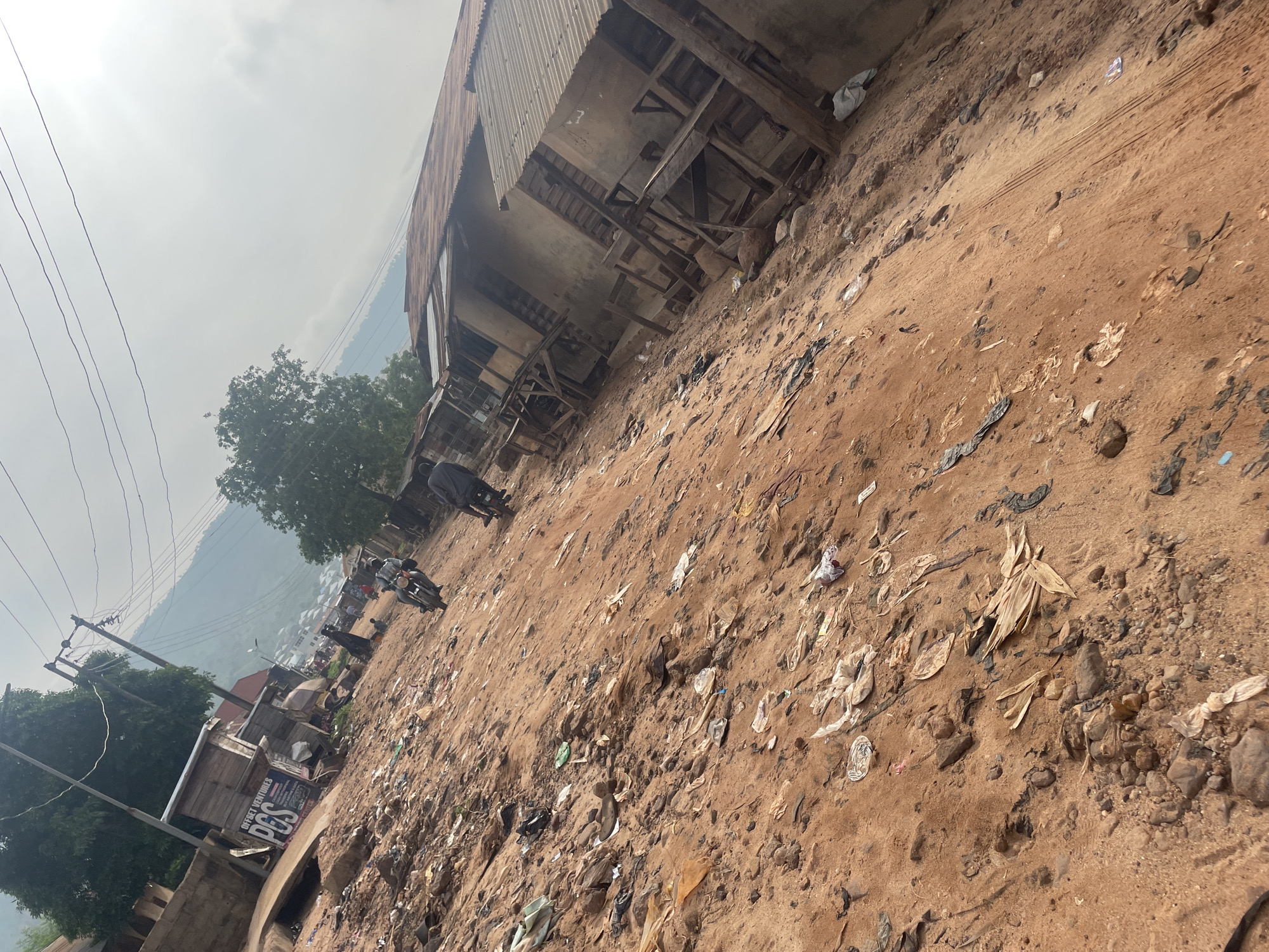 In Lokoja, Nigeria, a dirt road is littered with debris, including plastic bags, paper, and other refuse. The road is lined with simple structures constructed from wood and corrugated metal. A few people are visible, some on motorcycles and others walking. In the background, a hillside dotted with buildings and vegetation is visible under a hazy sky. The scene suggests a semi-urban or rural environment with an accumulation of waste.