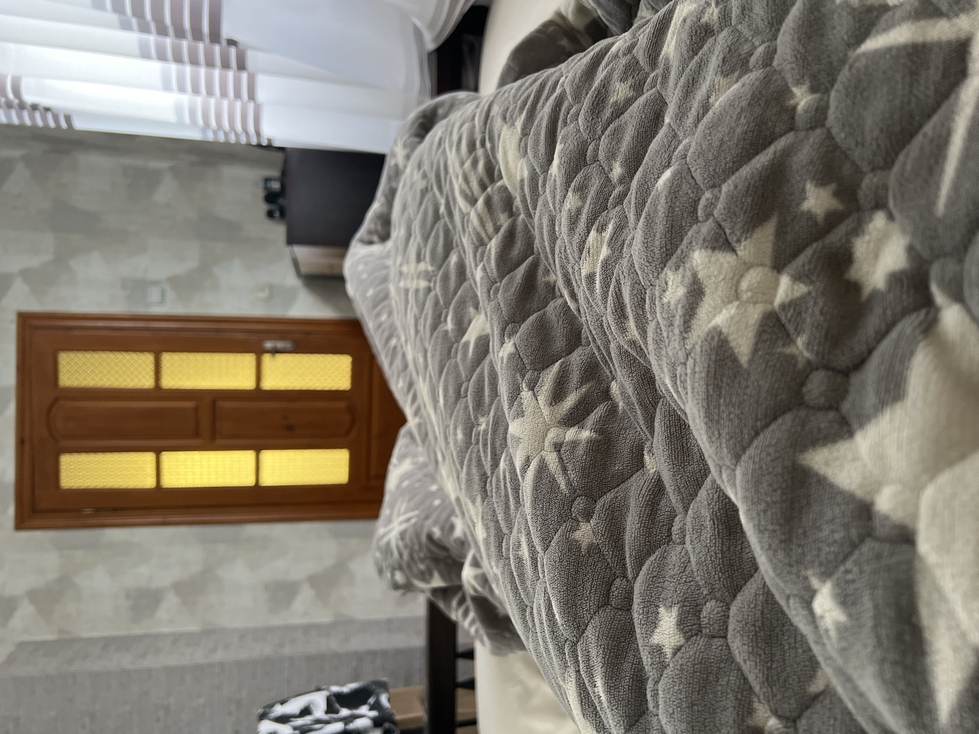 In a bedroom in Sobishchytsi, Ukraine, a person is covered by a grey blanket with white star patterns. The blanket is the main focus of the image, filling the foreground and extending into the background. Behind the bed, a wooden door with frosted glass panels, illuminated with a warm yellow light, stands partially open. To the right of the door, sheer white curtains with horizontal brown stripes are visible, suggesting a window. The walls appear to be a light, textured grey. A dark dresser or cabinet is partially visible to the right of the door, with a few objects on top. A dark wooden bed frame is also visible on the left side of the image. The overall impression is of a domestic, possibly morning or evening, scene in a residential setting.