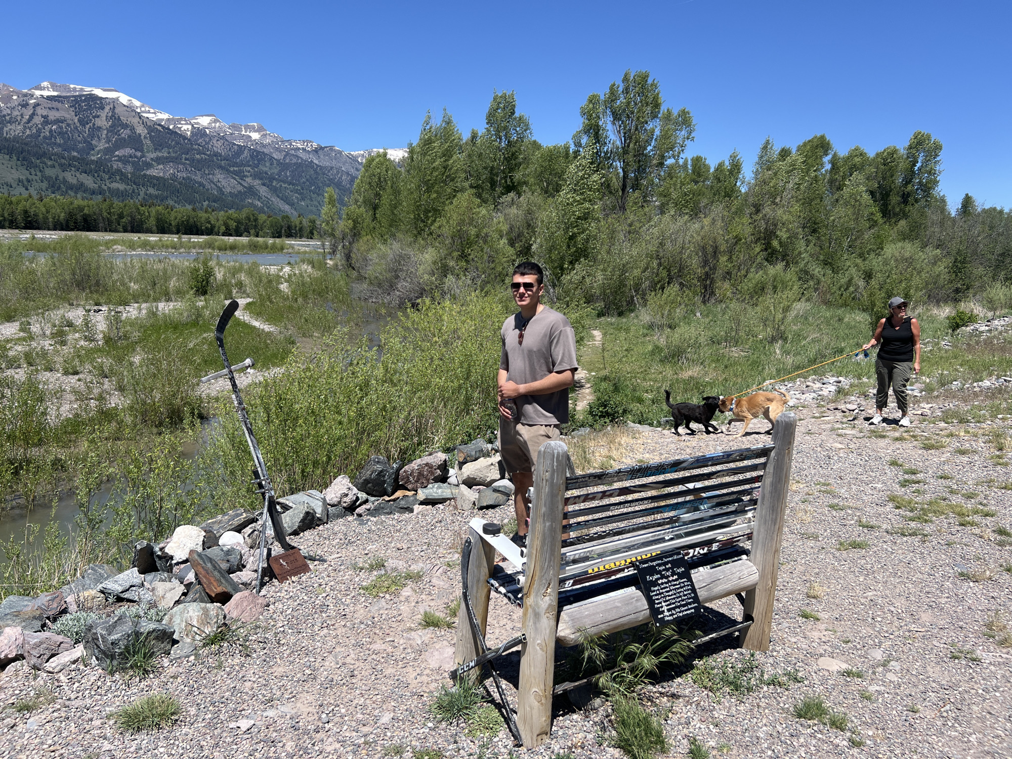 This detailed description pertains to an outdoor scene in Wilson, United States of America, captured on a clear, sunny day.
The image features two individuals and two dogs on a light-colored gravel path that runs along a body of water. In the foreground, slightly left of center, a fair-skinned man in sunglasses, a short-sleeved grey t-shirt, and khaki shorts stands, holding a clear plastic water bottle in his left hand and looking towards the viewer with a slight smile. To his immediate right, a rustic wooden bench with a seat and backrest constructed from numerous hockey sticks is visible. A black hockey puck rests on the left armrest of the bench, and a dark plaque with white text, including words like "Keylan," "Tiejie," "laughter," and "joy," is affixed below the seat, indicating a memorial. To the left of the bench, near the water's edge, an upright hockey stick is planted within a cluster of multi-colored rocks, at the base of which a small, dark rectangular plaque clearly displays the name "TJESIE."
Further along the gravel path on the right side of the image, a fair-skinned woman, wearing a dark cap, a black tank top, and dark pants, walks away from the viewer, looking slightly to her left. She holds a yellow leash connected to a medium-sized tan dog. A smaller black dog is also present on the path, interacting with the tan dog.
The environment includes a calm, light blue-grey river or creek flowing across the middle ground. The foreground and riverbanks are covered in lush green shrubs and grasses. A dense band of green trees lines the far bank of the water body. In the background, majestic snow-capped mountains rise under a clear, cloudless blue sky, their lower slopes covered with a dark green treeline. The overall scene suggests a leisurely outdoor setting with memorial elements.