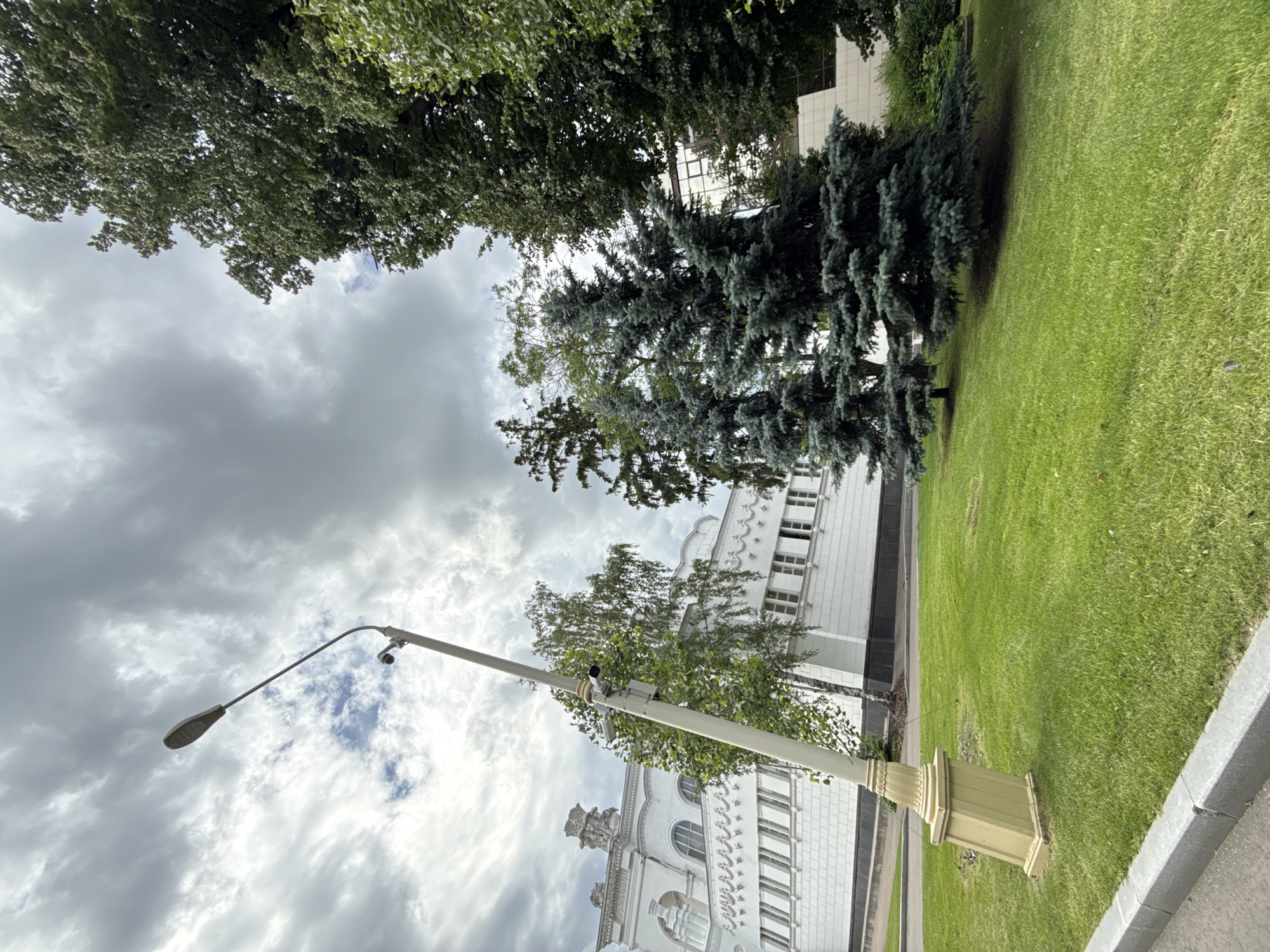 The image shows an outdoor scene with a building, trees, a lamp post, and a grassy area. The building is white with many windows and decorative architectural features. Two large trees, one with dark green needles and another with lighter green leaves, stand in front of the building. A tall lamp post with a curved arm and a light fixture is visible in the foreground. There are also what appear to be security cameras mounted on the lamp post. The ground is covered with a well-maintained green lawn. The sky is overcast with dramatic clouds. The image is oriented vertically. The overall impression is of a well-kept urban or park area. The time of day appears to be daytime, given the lighting. The weather is cloudy. The location is specified as Moscow, Russia, which might suggest the architectural style of the building or the types of trees. There is no visible text in the image.