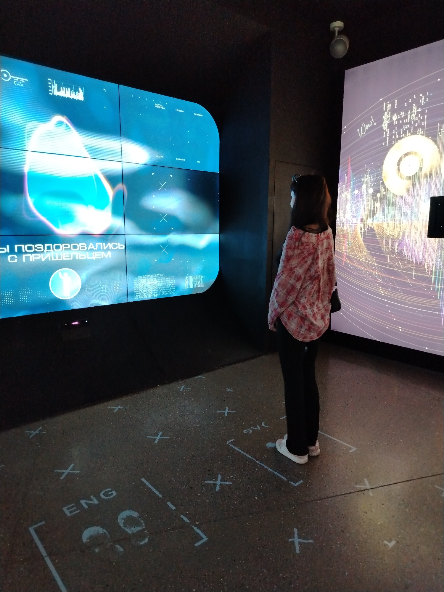 The image shows a woman standing in front of two large screens. The screen on the left displays abstract blue and white imagery with text in Russian that appears to read "ВЫ ПОЗДОРОВАЛИСЬ С ПРИШЕЛЬЦЕМ" which translates to "YOU CONGRATULATED THE ALIEN". The screen on the right shows a colorful, abstract graphic with the word "Wow!" written in script. The floor in front of the screens has a pattern of white lines and crosses, with text labels like "ENG". This setup suggests an interactive exhibit, possibly in a museum or public space. The lighting is dim, with the screens being the primary light sources. The woman is wearing a plaid shirt and dark pants. The overall environment is modern and dark. The specific location in Moscow, Russia cannot be determined from the image alone.
