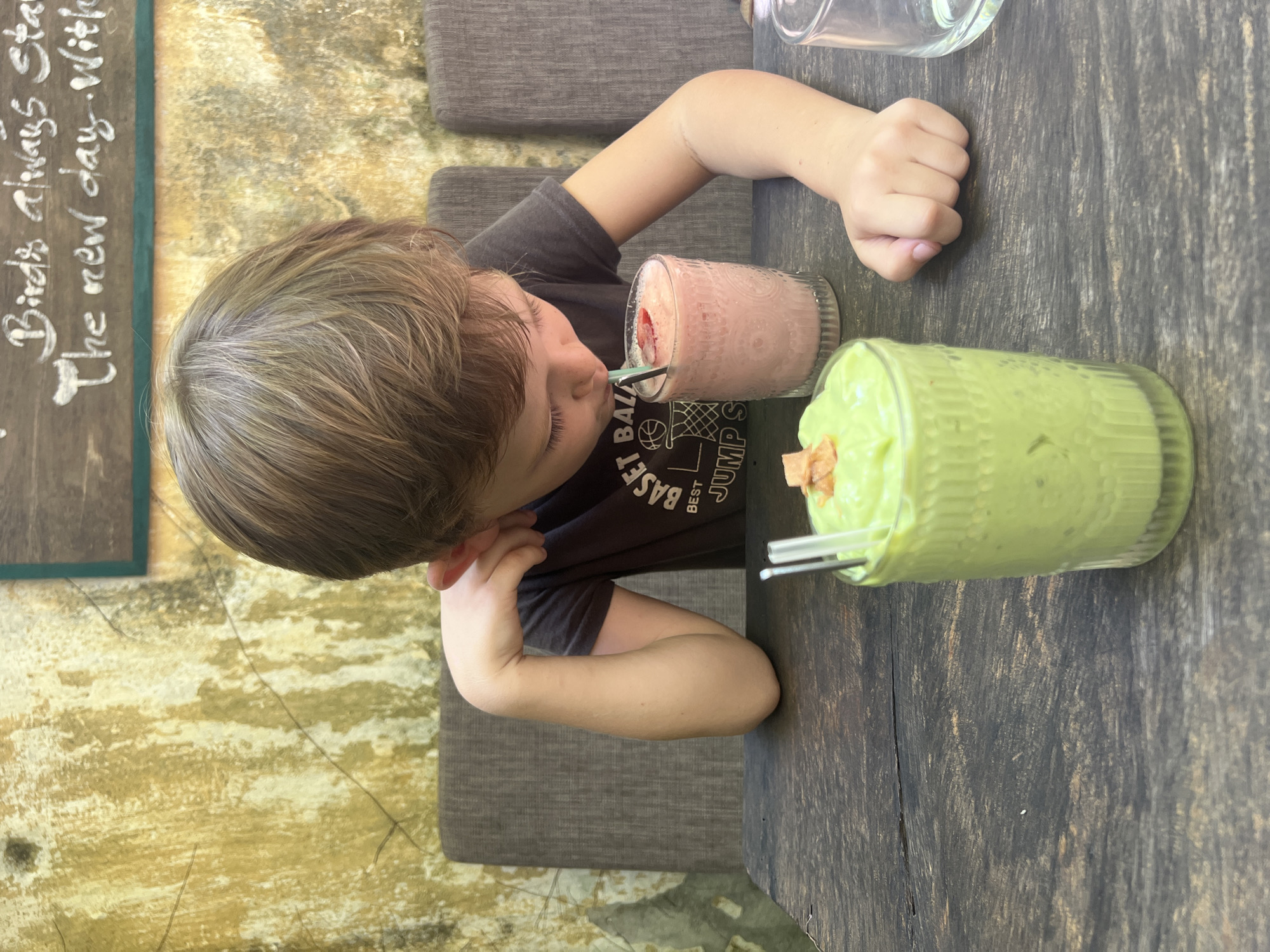 A young boy with brown hair is seated at a dark wooden table, leaning forward and drinking from a pink smoothie with a blue straw. He is wearing a dark t-shirt with a graphic print that includes the words "BEST BALL" and "JUMP S". To his left, another glass contains a green smoothie topped with small pieces of what appears to be fried bread or crackers. The background features a textured, yellowish-green wall with faint dark lines, reminiscent of aged plaster or paint. Above the boy's head, a wooden board with white chalk writing is visible, part of which reads "Birds always star The new day. With". The setting suggests an outdoor or cafe environment in Hội An Ward, Vietnam, possibly a restaurant with a rustic or artistic theme. The lighting appears to be natural daylight, casting soft shadows. The boy's left elbow rests on the table while his right hand is near the green smoothie. Two chairs, upholstered in a dark grey fabric, are visible behind the boy.